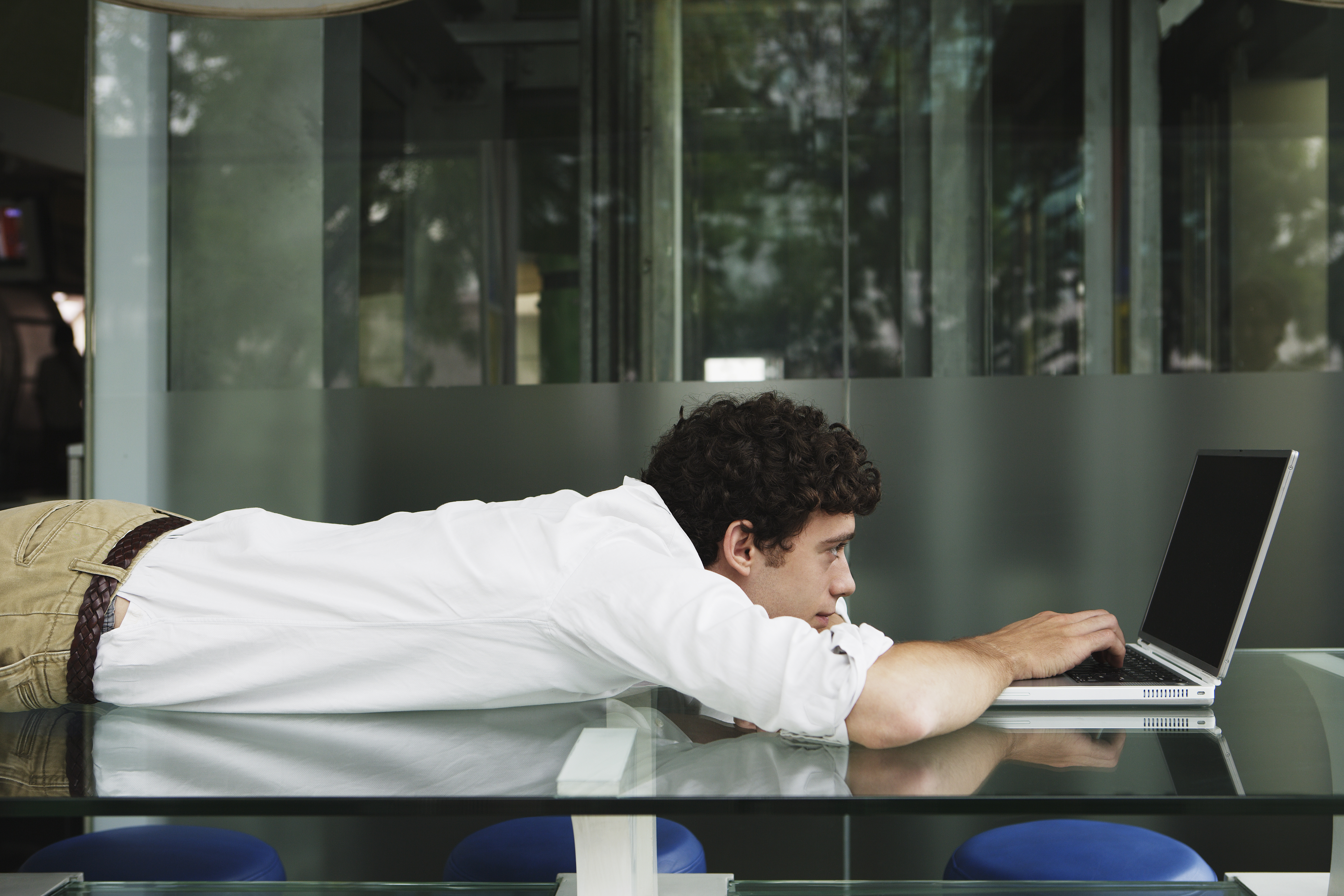 Young man lying on glass table using laptop computer, side view