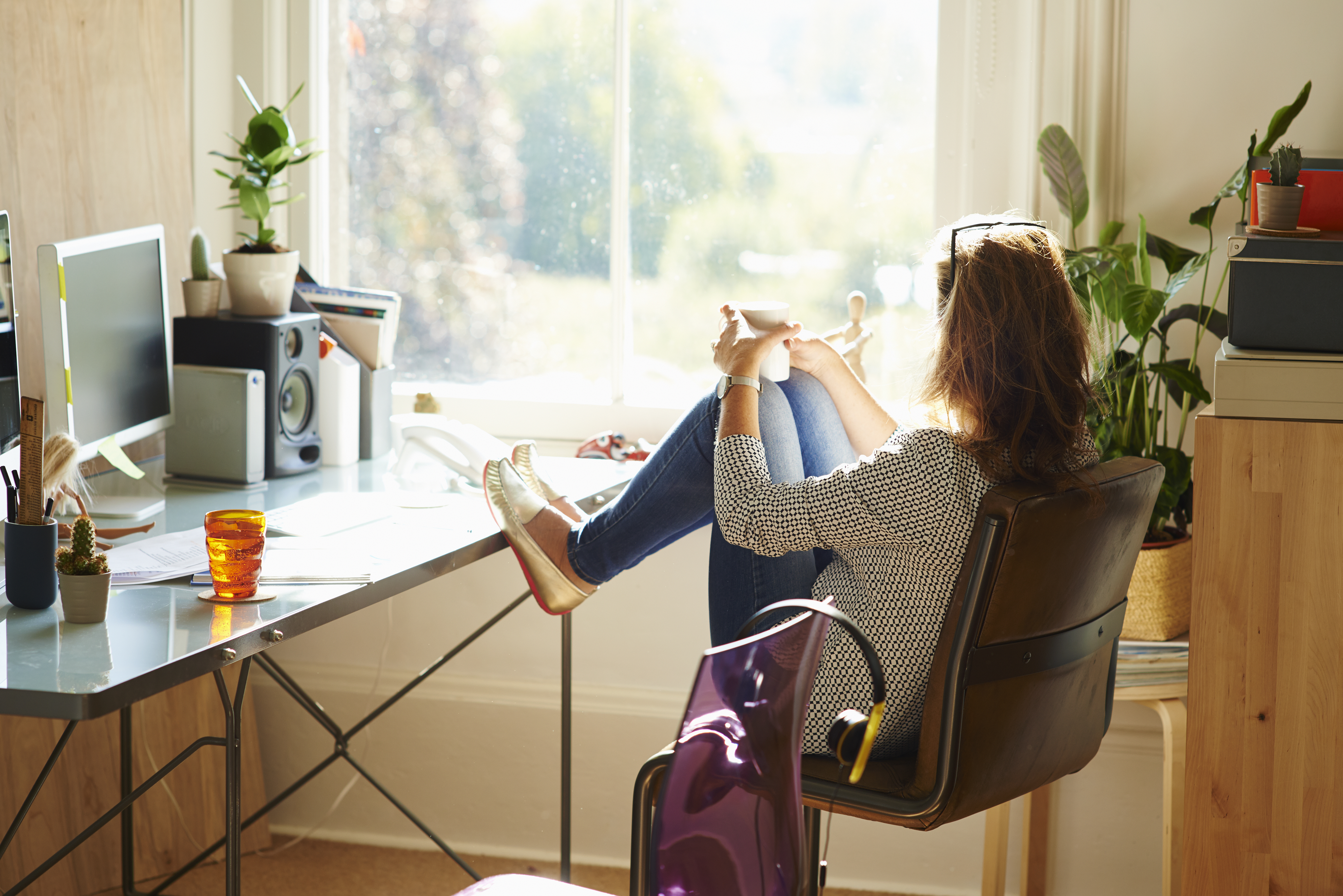 Pensive woman looking through window with feet up on desk in sunny home office