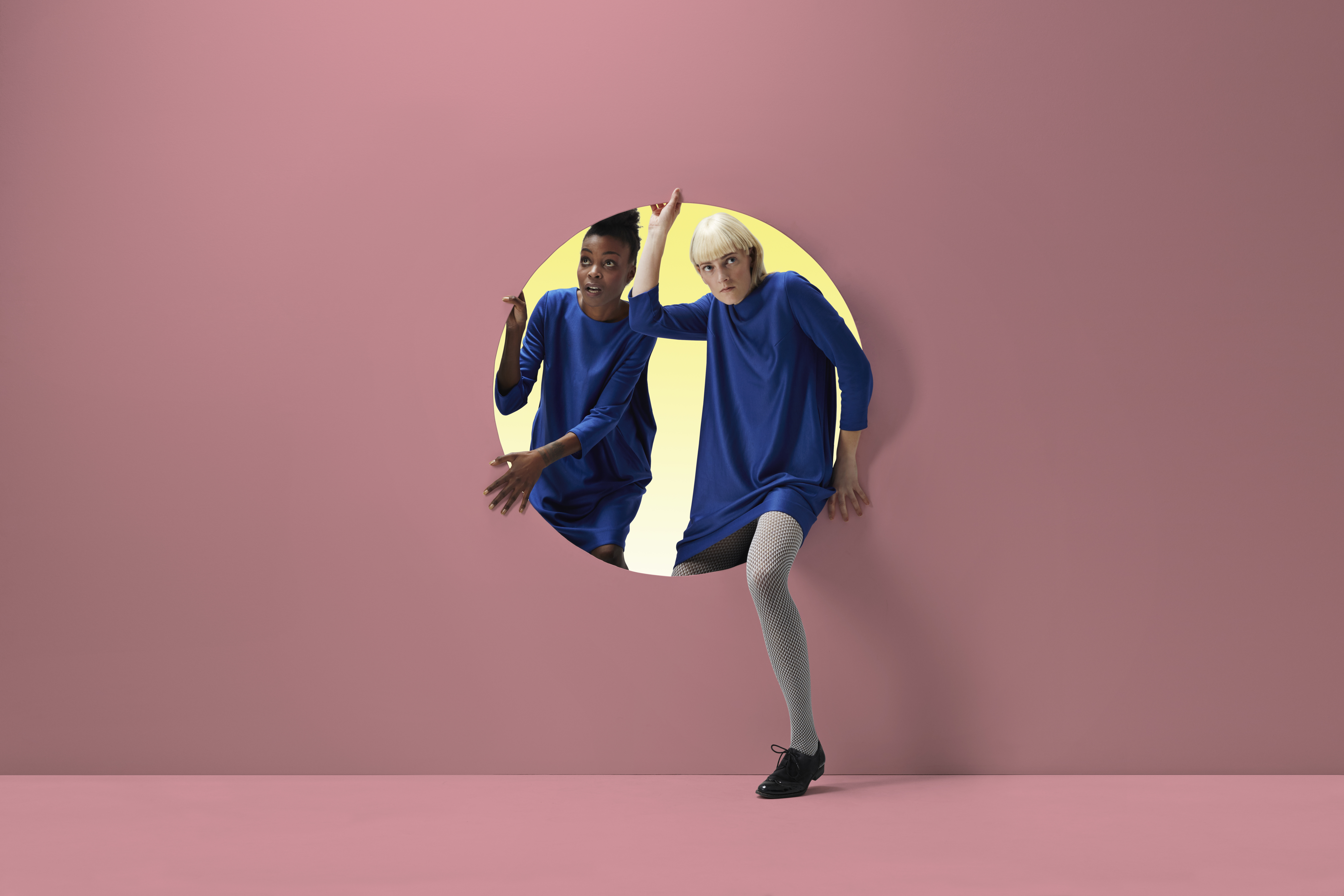Two women peeking out of round opening in coloured wall