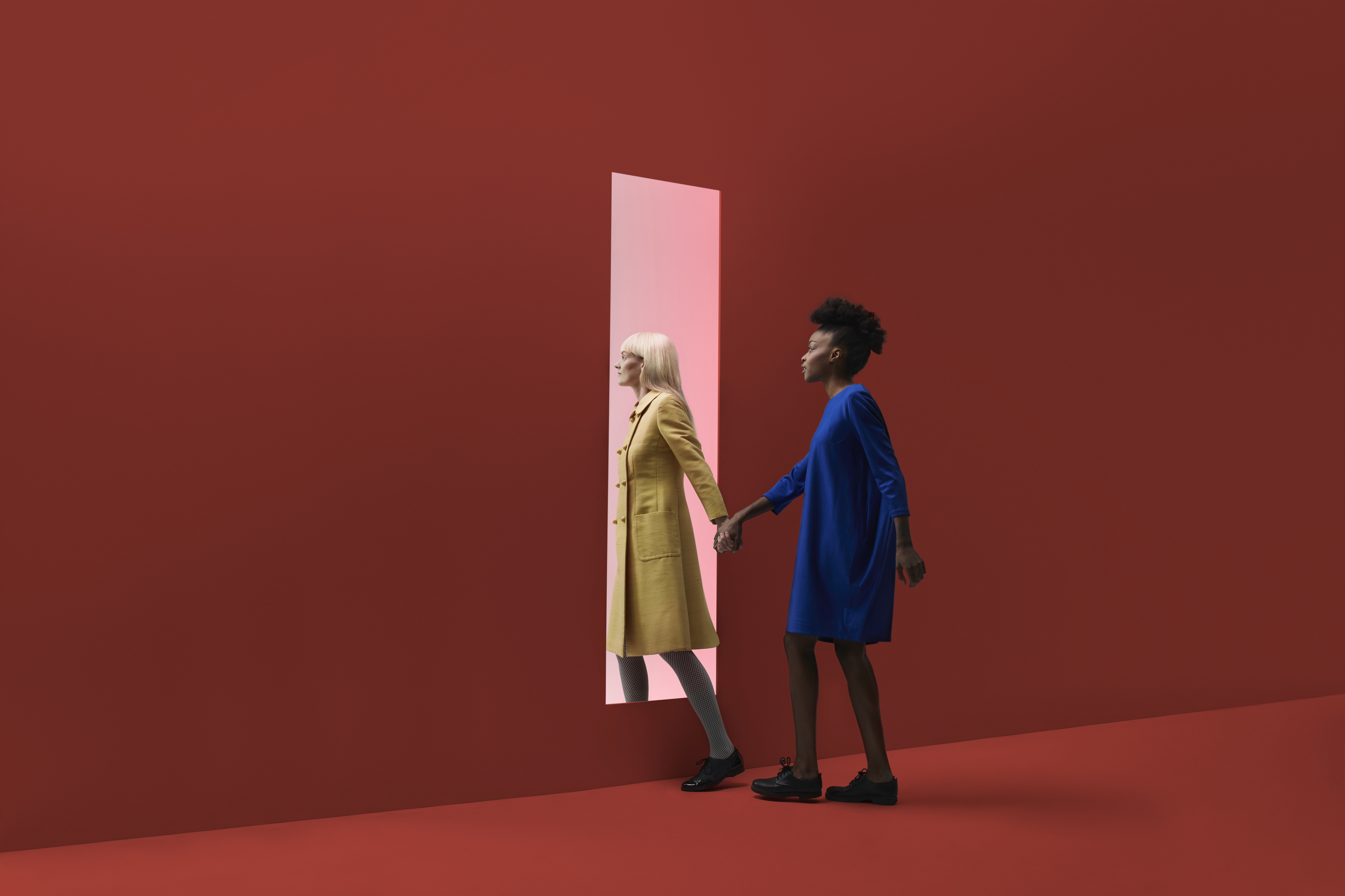Two women holding hands, walking threw rectangular opening in coloured wall