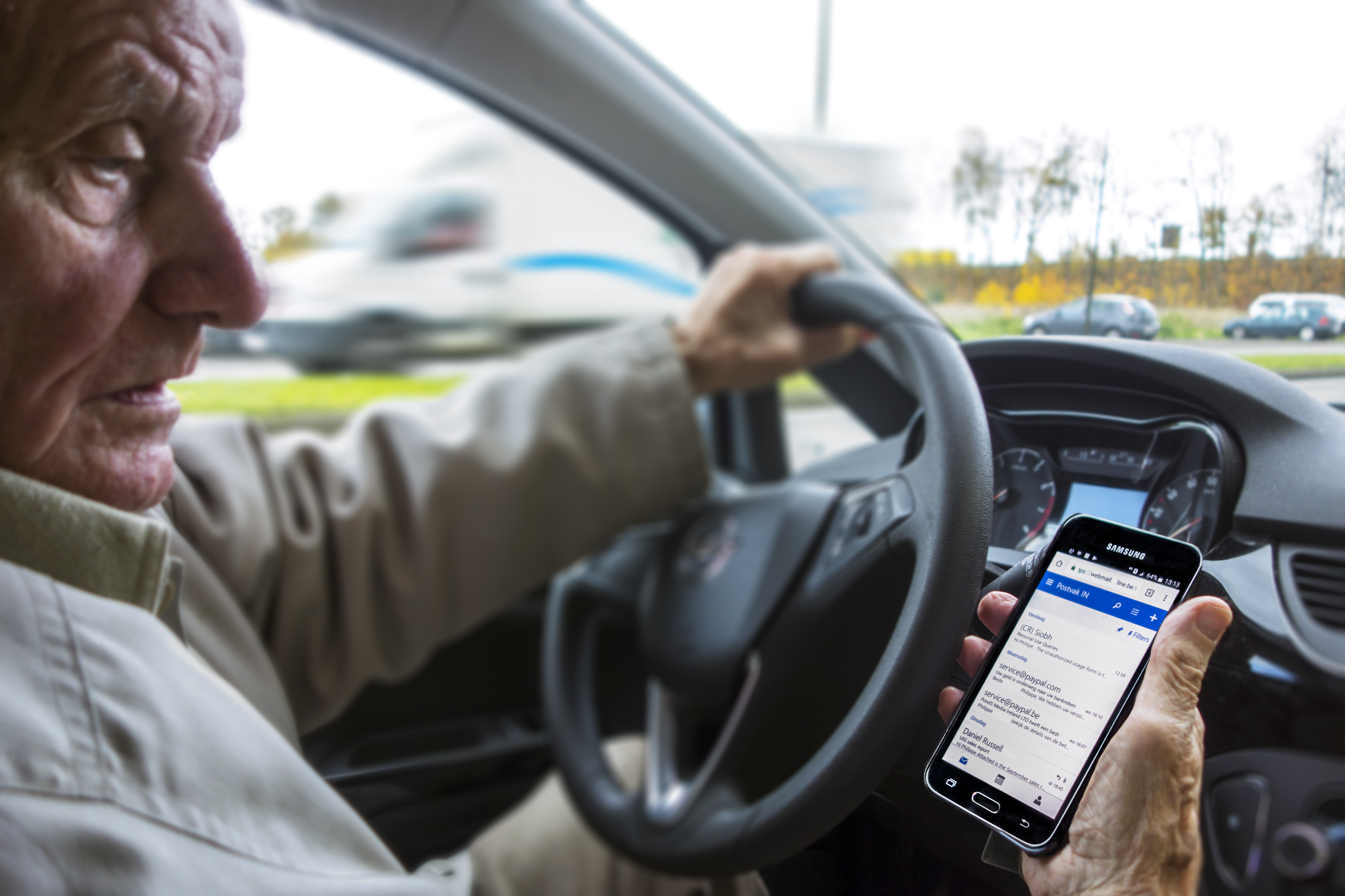 Elderly man at steering wheel checking messages on smart phone while driving car on road.