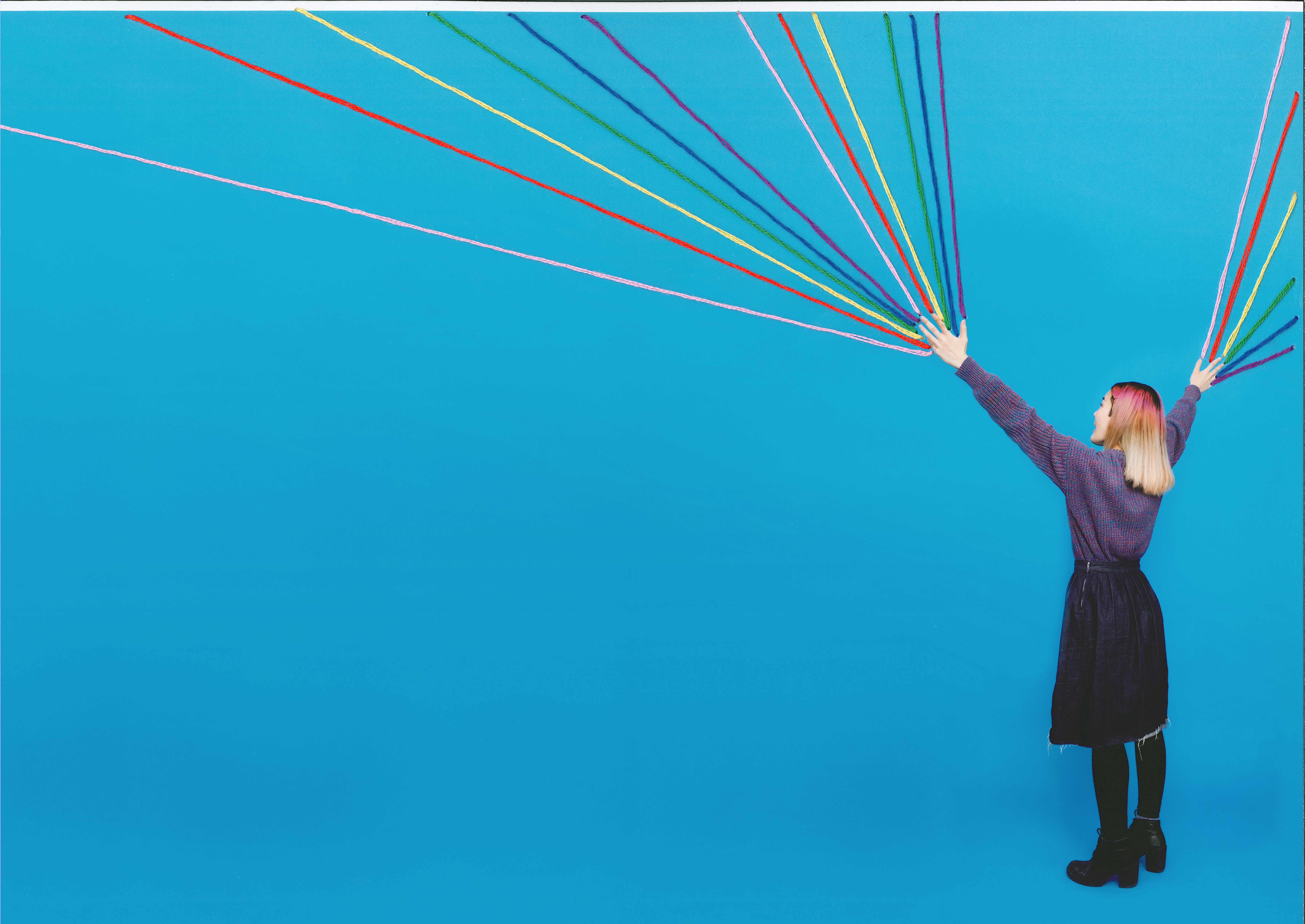 A woman faces away from the camera. She has her arms raised and rainbow strands shoot out of her fingertips.