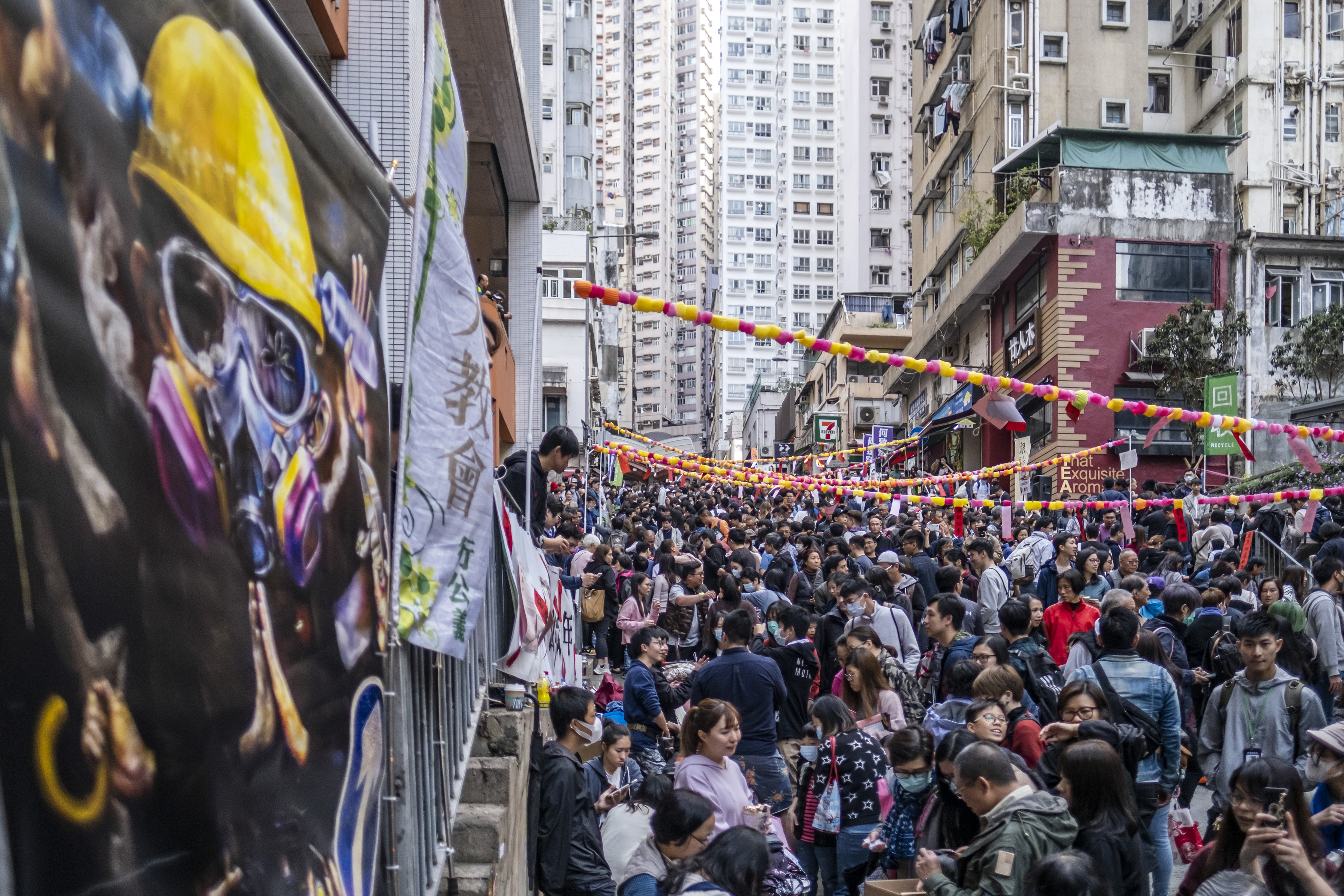People walk past a protest banner during a Lunar New Year fair organized by pro-democracy district councilors on Centre Street in the Sai Ying Pun district of Hong Kong, China, on Saturday, Jan. 18, 2020. The Lunar New Year, when spending traditionally peaks in Hong Kong amid a flood of vacationing Chinese mainlanders, will likely not provide its usual boost after months of fiery demonstrations that have driven tourists away. Photographer: Justin Chin/Bloomberg via Getty Images