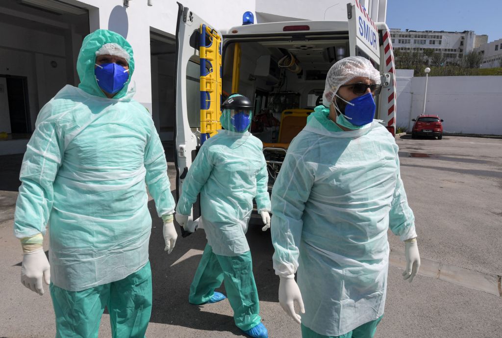 Three doctors in protective equipment walking outside with an ambulance in the background.