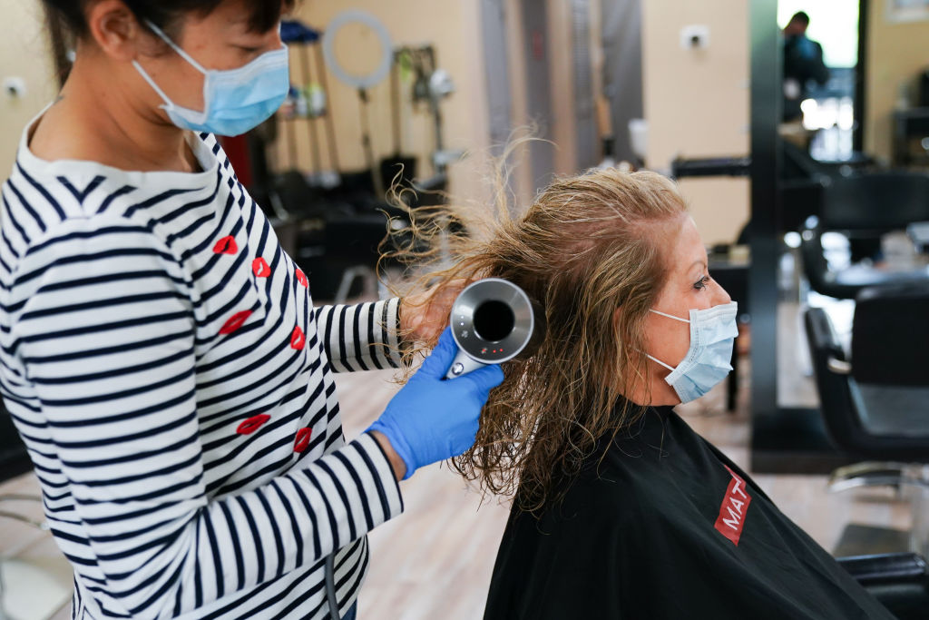 Hairdresser in face mask blow drying the hair of a woman who also wears a face mask.