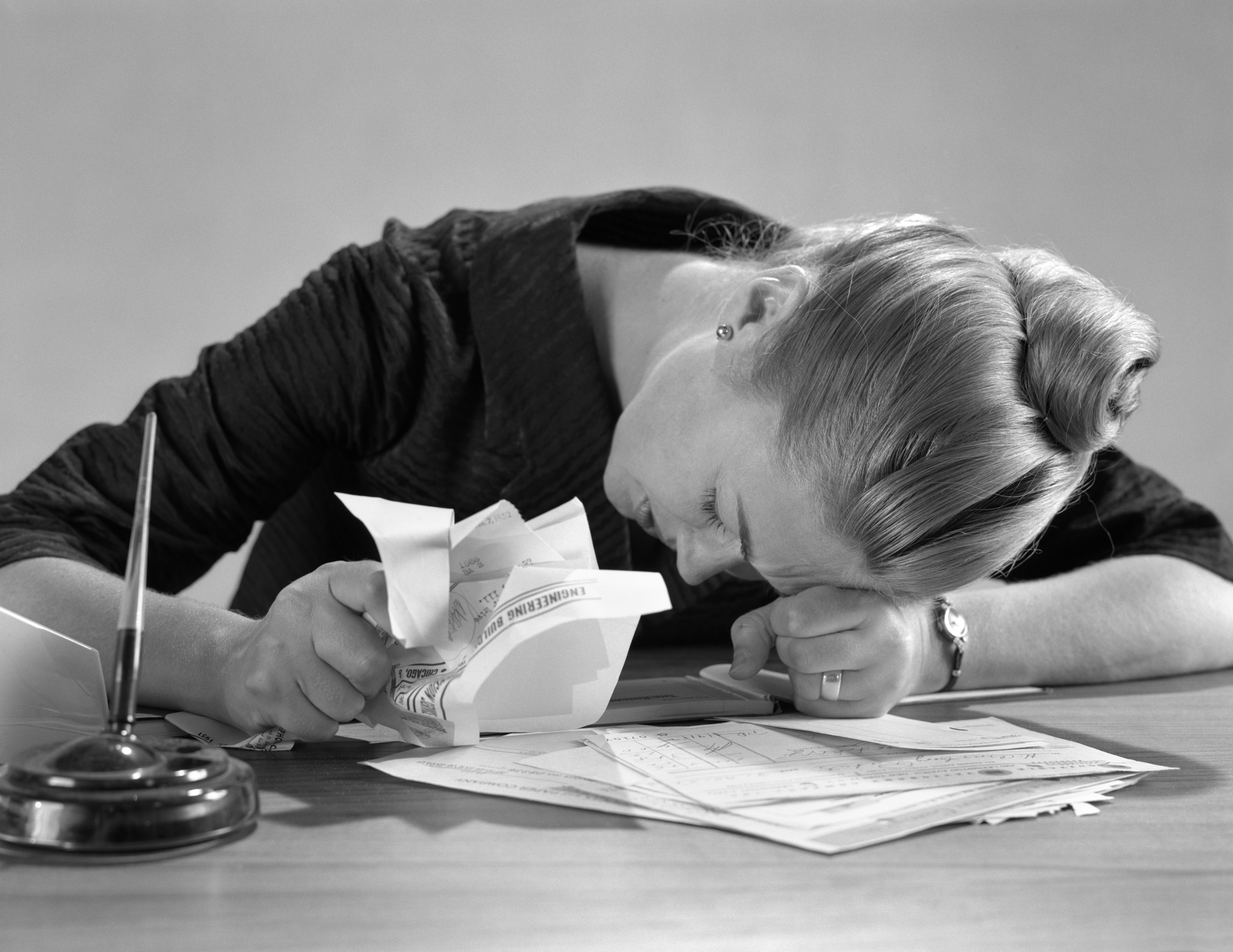 1960s UNHAPPY WOMAN SLUMPED ACROSS DESK OVER PAPERS BILLS GRASPING SOME IN HAND (Photo by H. Armstrong Roberts/ClassicStock/Getty Images)