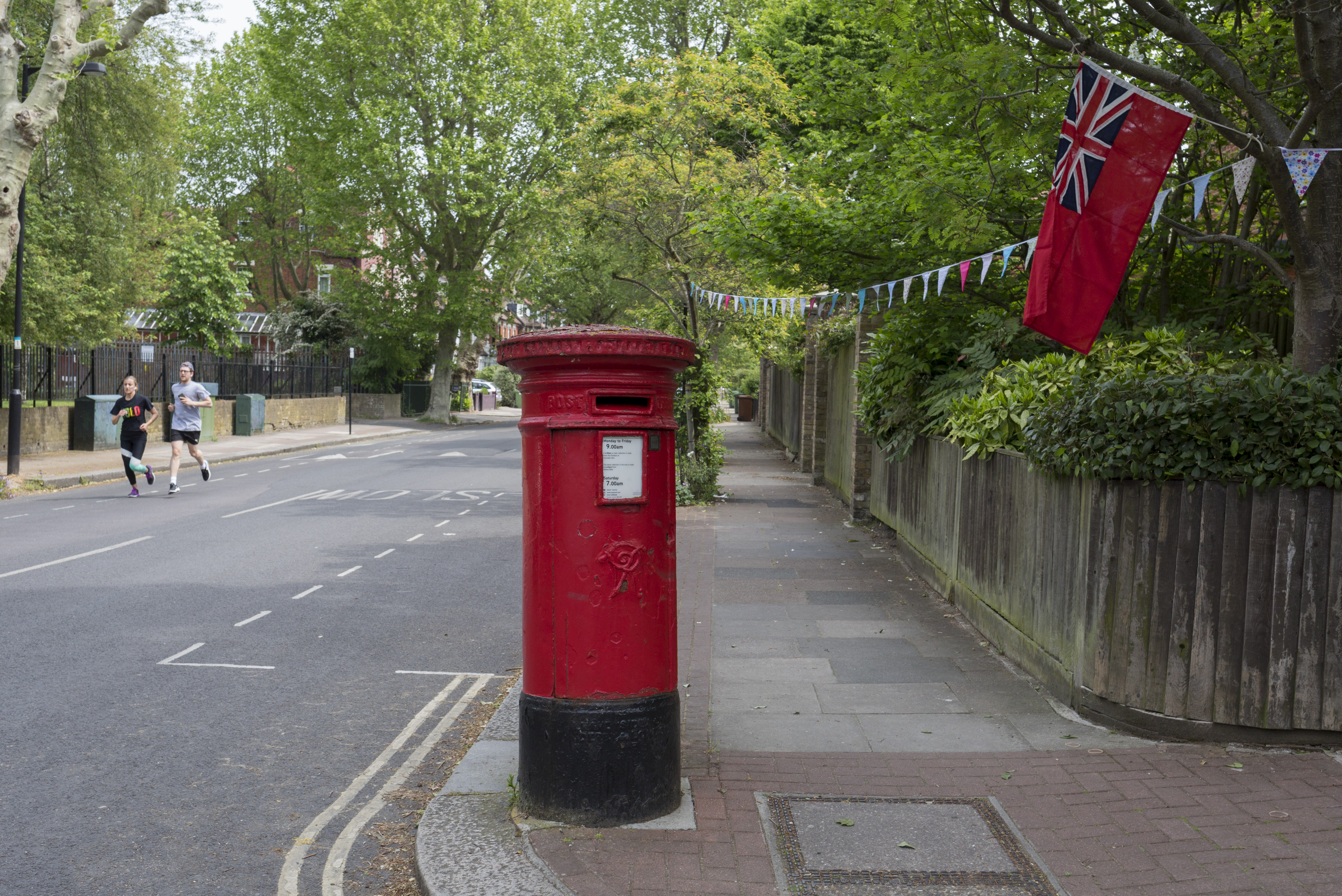On the 75th anniversary of VE Day (Victory in Europe Day, the official end of WW2) and during the UK's Coronavirus pandemic lockdown, two runners jog towards a heritage Royal Mail post box and a naval ensign flag that hangs from the garden of a corner property in Camberwell, on 8th May 2020, in London, England. (Photo by Richard Baker / In Pictures via Getty Images)