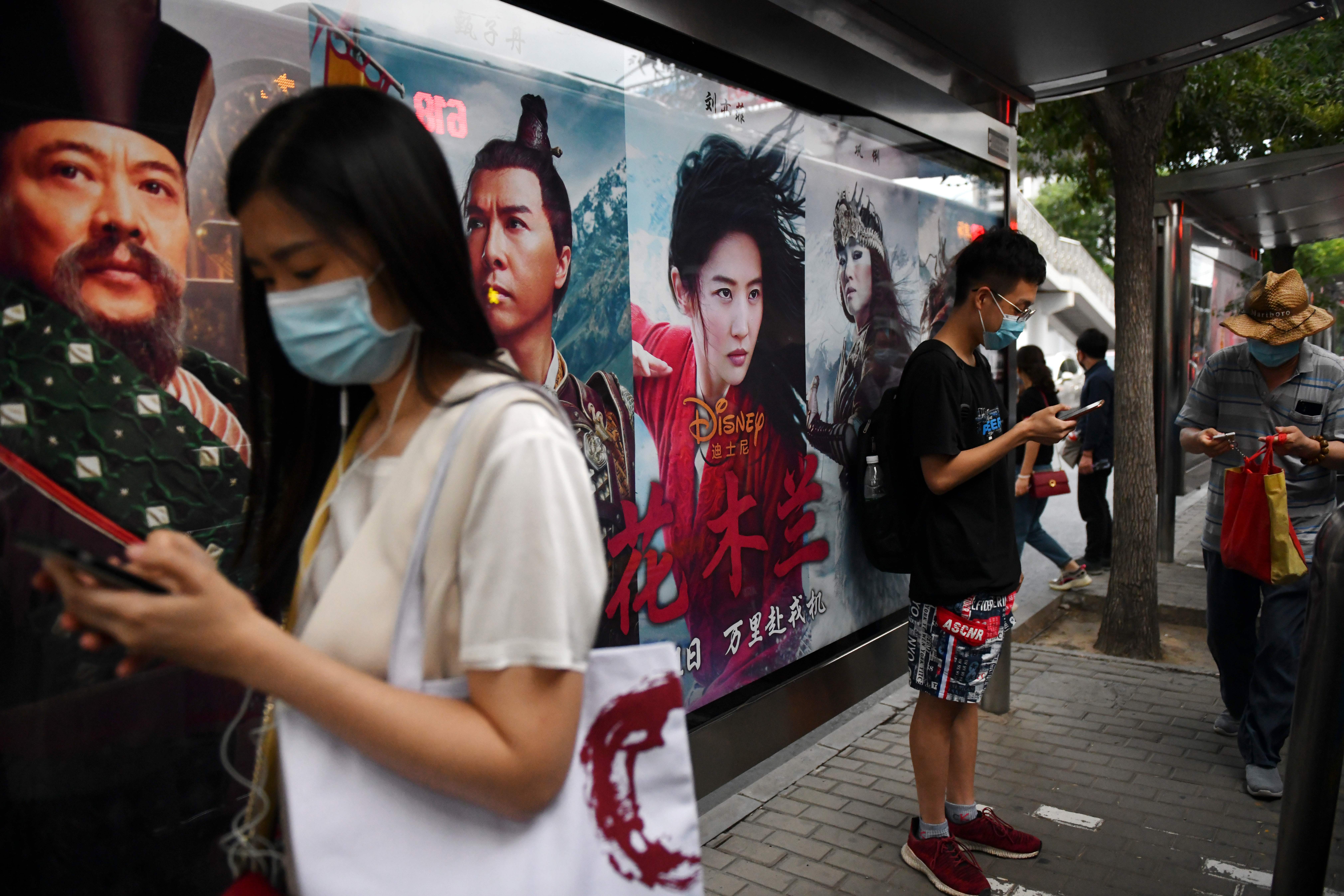 People wait beside a poster for the Disney movie "Mulan" at a bus stop on the day the movie opened in Beijing on September 11, 2020. - Pilloried internationally and given a lukewarm debut by Chinese cinemagoers, Disney on September 11 discovered its $200 million live-action epic "Mulan" has at least one ardent fan -- China's firebrand foreign ministry. (Photo by GREG BAKER / AFP) (Photo by GREG BAKER/AFP via Getty Images)
