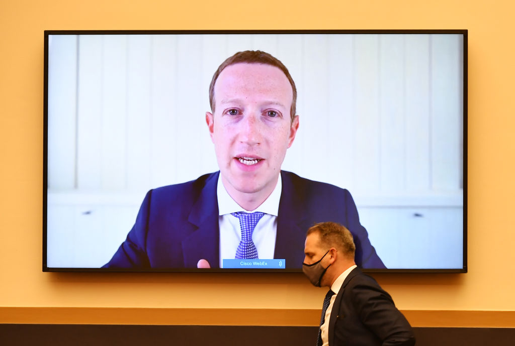 Photo of a big TV screen filled with an image of Facebook CEO Mark Zuckerberg testifying before Congress while a man in a suit and with a mask on walks in front of the screen.