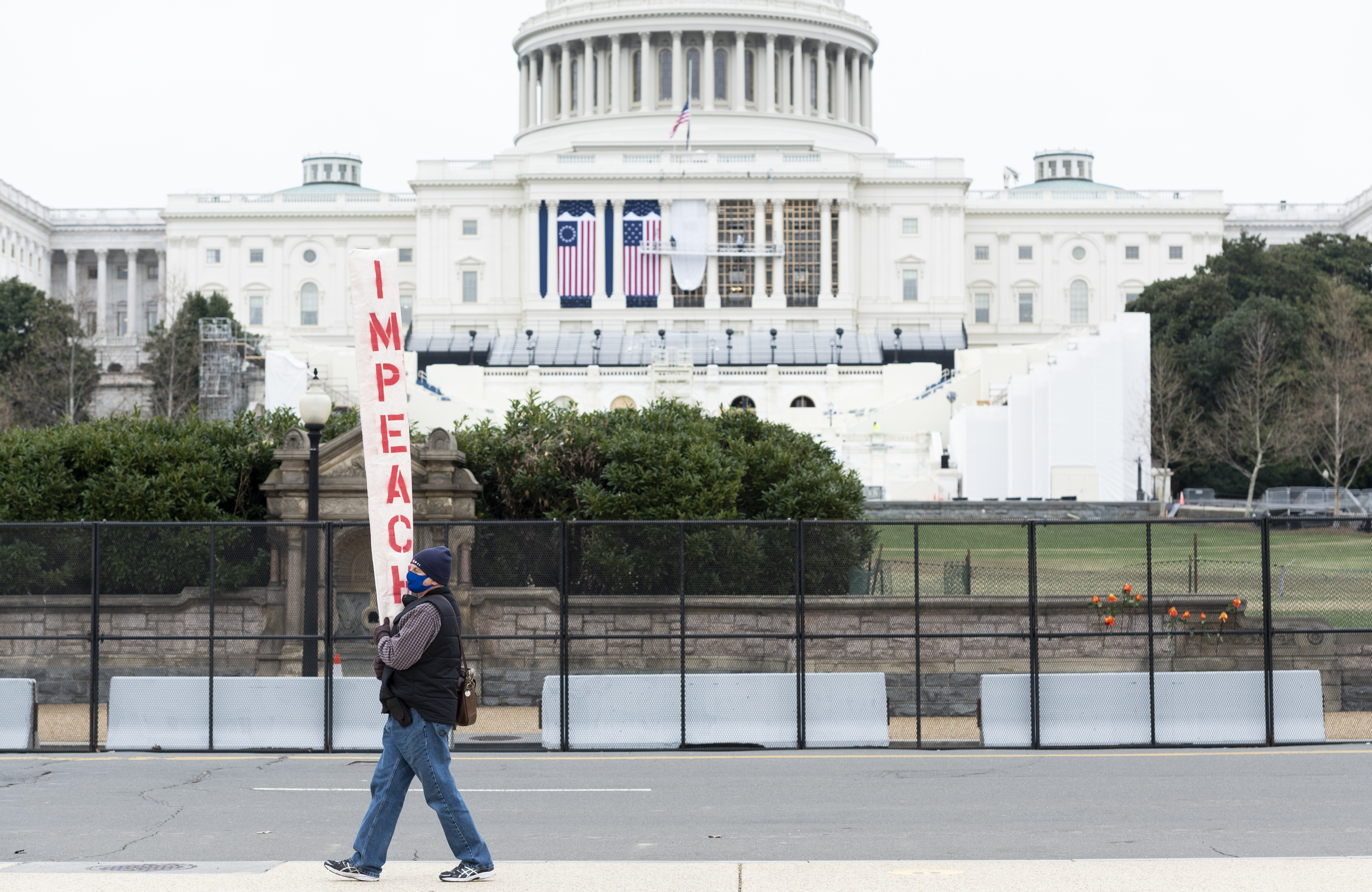 Workers hang the flags on the West Front facade for the inauguration as a man carries an impeach sign past the Capitol on Jan. 11, 2021.