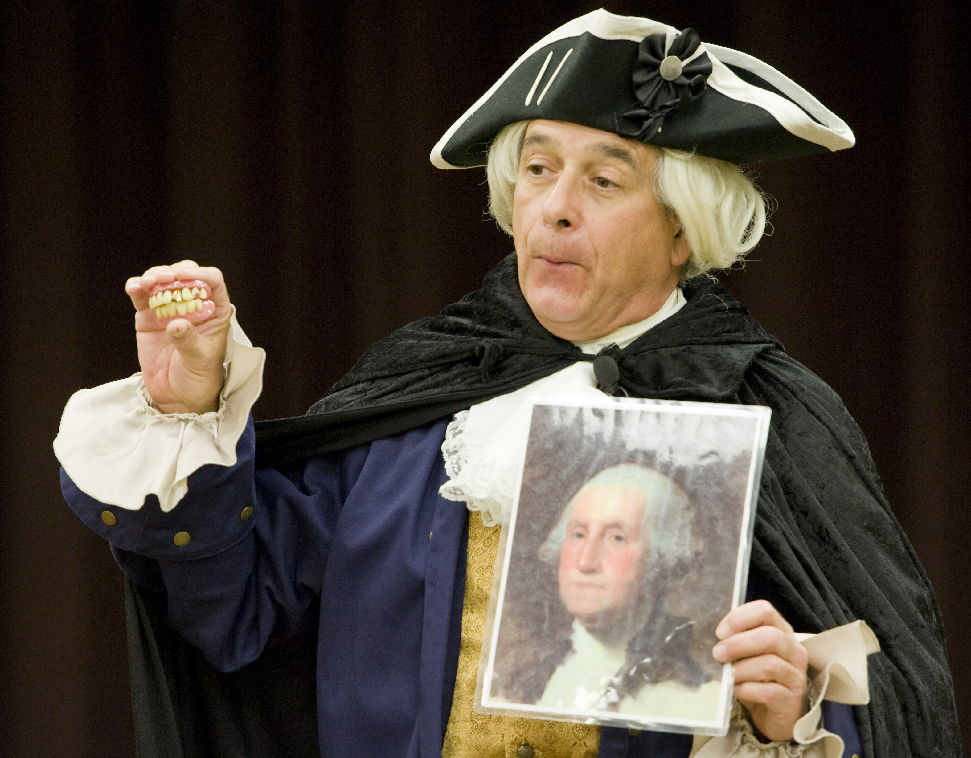 History educator Peter Small, portraying George Washington, holds a set of dentures and a portrait of Washington. Contrary to myth, Washington's dentures were made not of wood, but of an array of materials including gold, ivory - and the teeth of slaves.