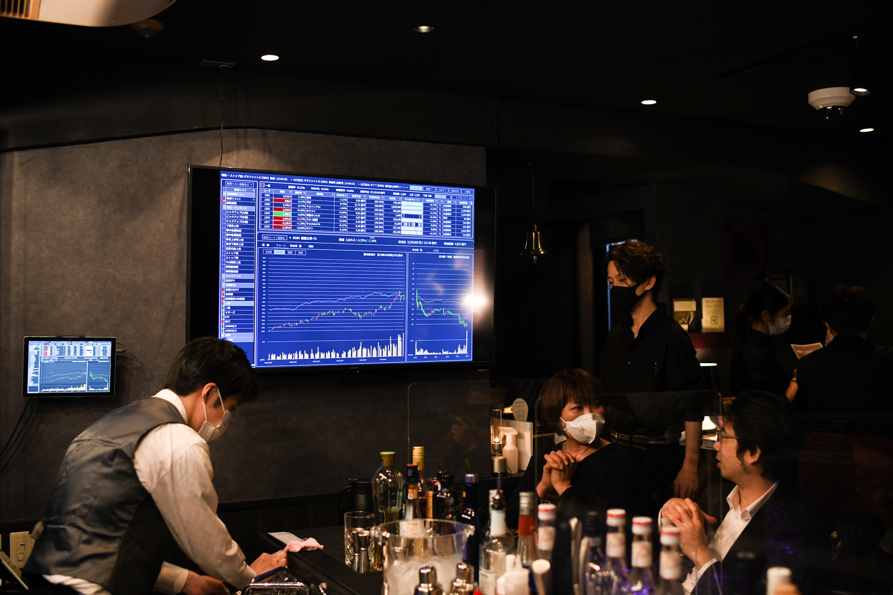 Customers next to a screen displaying financial information inside Stock Pickers, a tip-trading bar for investors, in the Ginza district of Tokyo, Japan, on Monday, March 29, 2021. Stock Pickers’ walls are decorated with books on value investing and how to bet like legendary investor Warren Buffett.