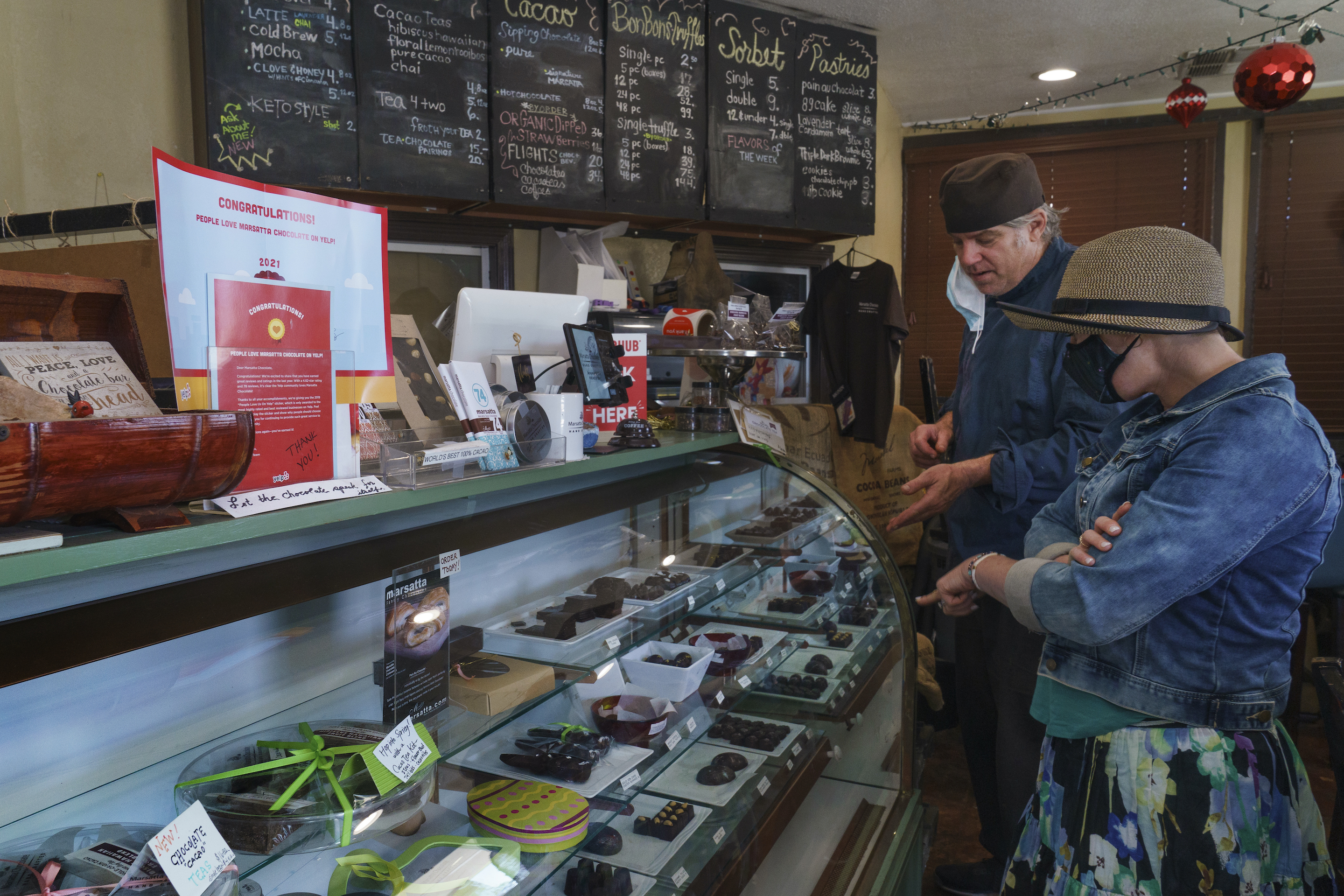 Jeffray Gardner, the owner of Marsatta Chocolate explains his artisan hand-crafted chocolates to new customer, Katherine Trontman at his company's flagship store in Torrance, Calif., Sunday, March 28, 2021. Restaurants and delivery companies remain uneasy partners, haggling over fees and struggling to make the service profitable for themselves and each other. Gardner says he probably loses money on the one or two delivery orders he gets each day. But he’s still happy to work with delivery companies because they help him reach a wider audience.