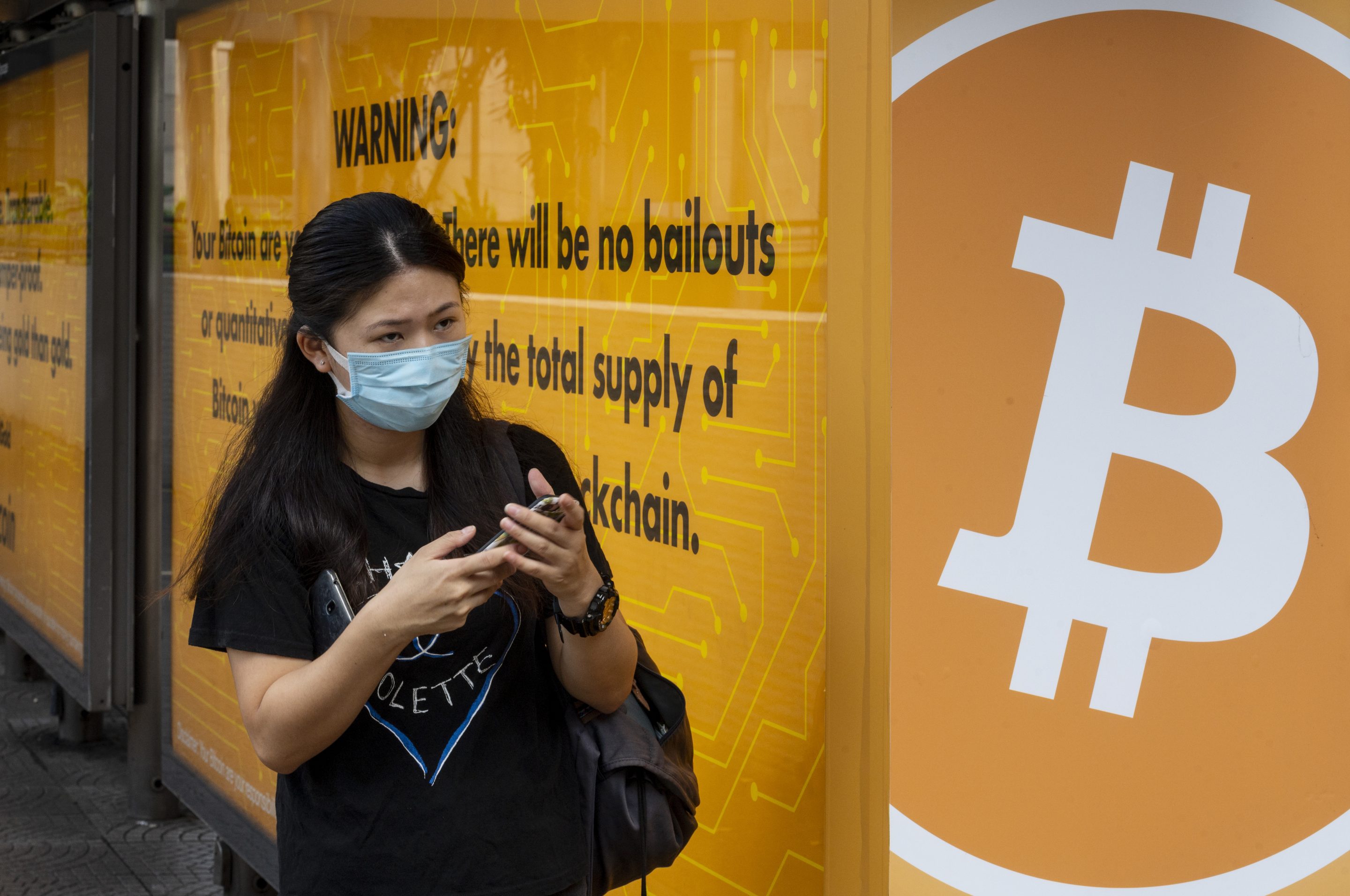 A woman wearing a mask stands next to a bus stop with a Bitcoin advertisement in Hong Kong.