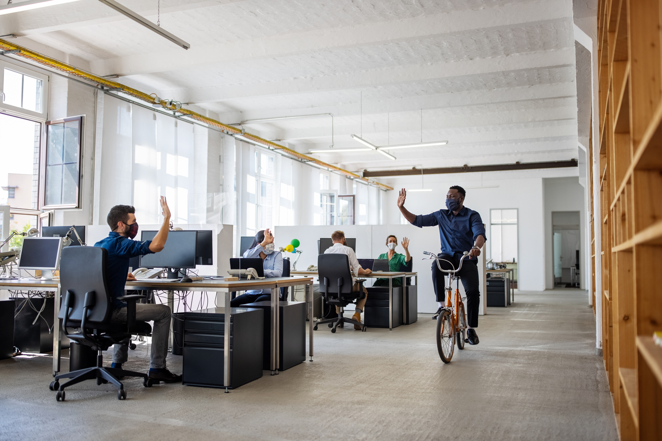 A businessman waving a hand to colleagues while riding a bicycle in a creative office. All employees are wearing face masks while working.