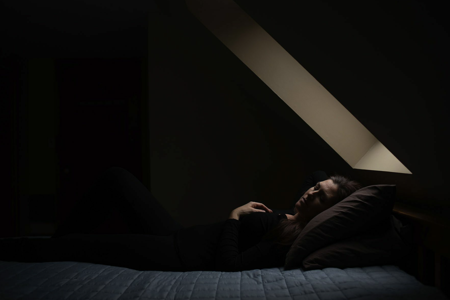 Woman laying on bed with eyes closed in dark room under a sky light.