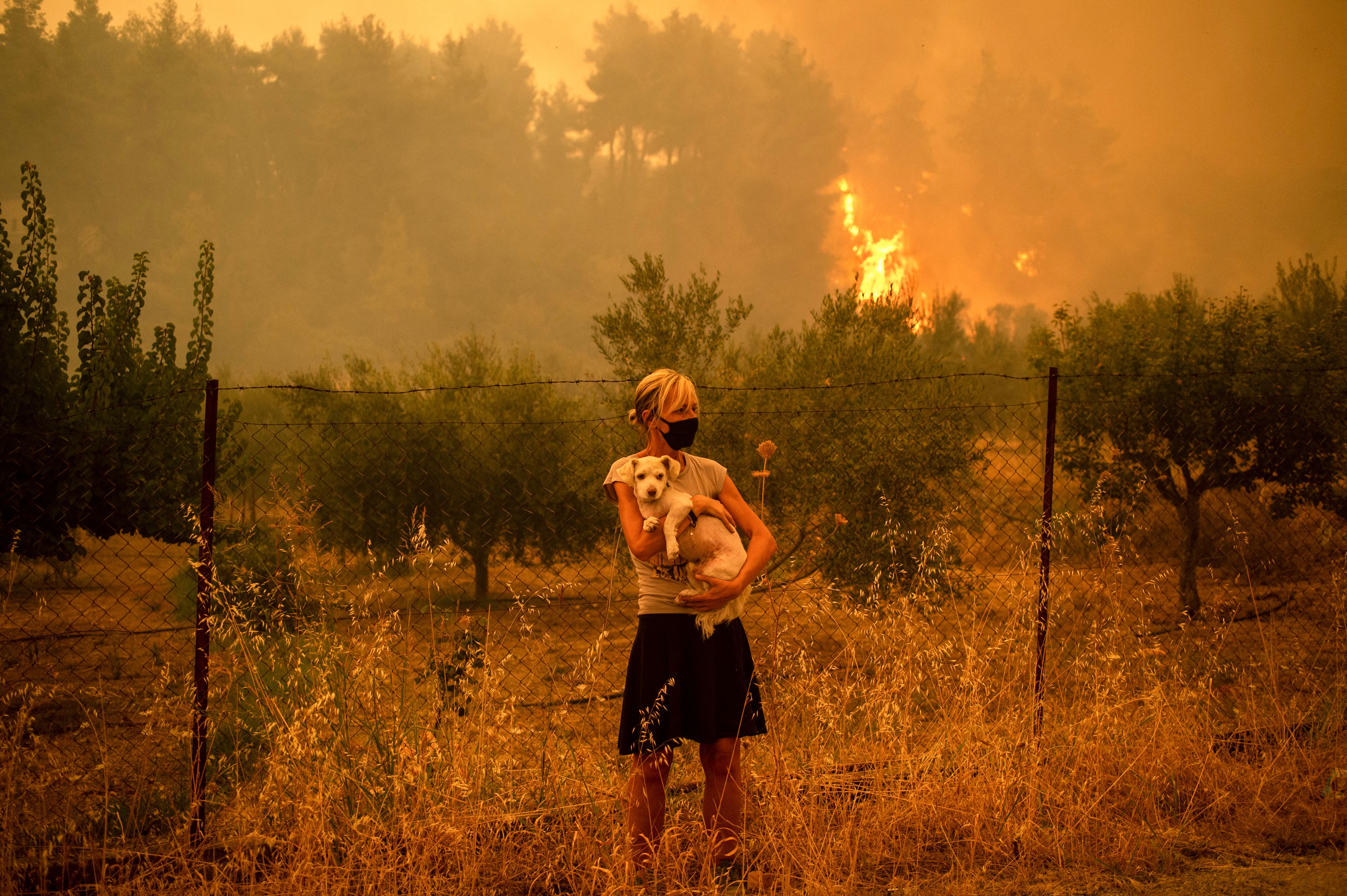 A woman holds a dog in her arms as forest fires approach the village of Pefki on Evia (Euboea) island, Greece's second largest island, on August 8, 2021. - Hundreds of Greek firefighters fought desperately on August 8 to control wildfires on the island of Evia that have charred vast areas of pine forest, destroyed homes and forced tourists and locals to flee. 