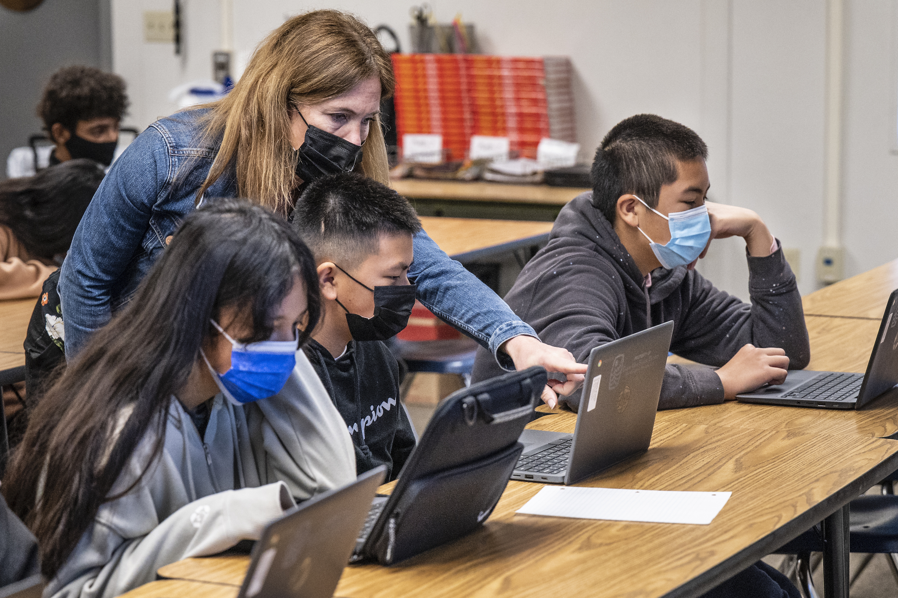 A teacher and three students wearing masks working on laptops in a classroom