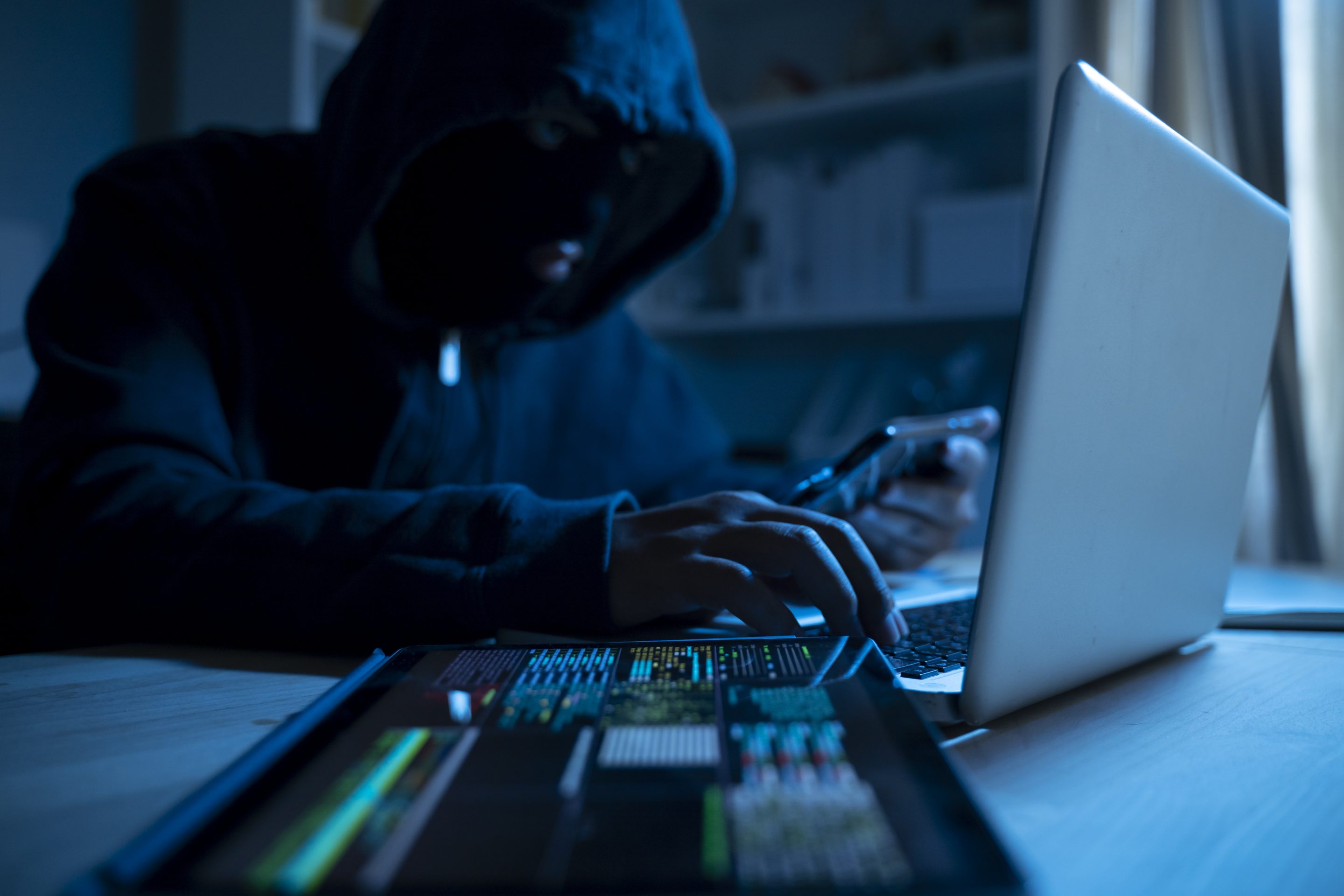 Anonymous person in the hood sitting in front of computer working with laptop and mobile phone