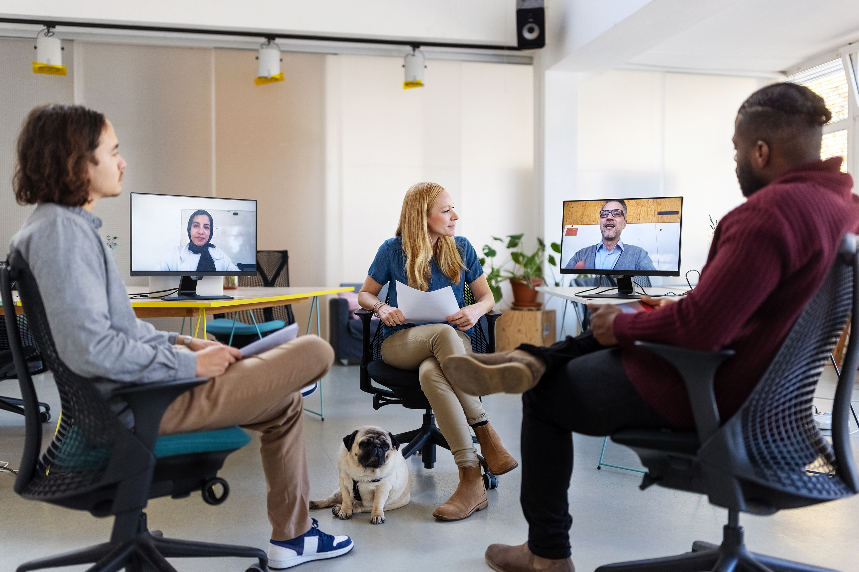 Team in office sitting in a room during a hybrid meeting with two remote employees.