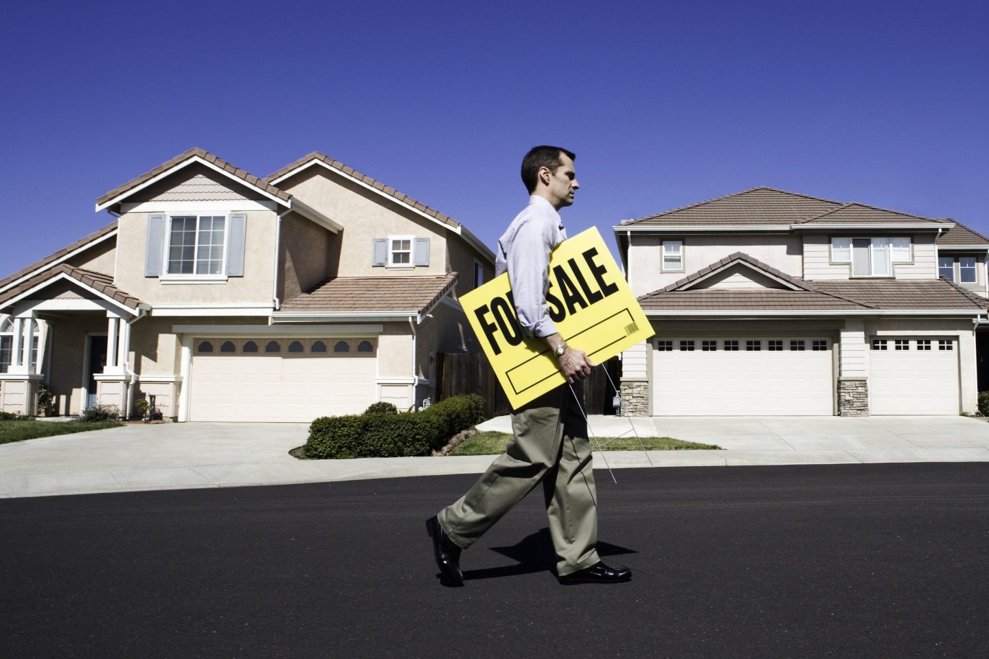 Male estate agent walking in front of house with holding for sale sign, side view