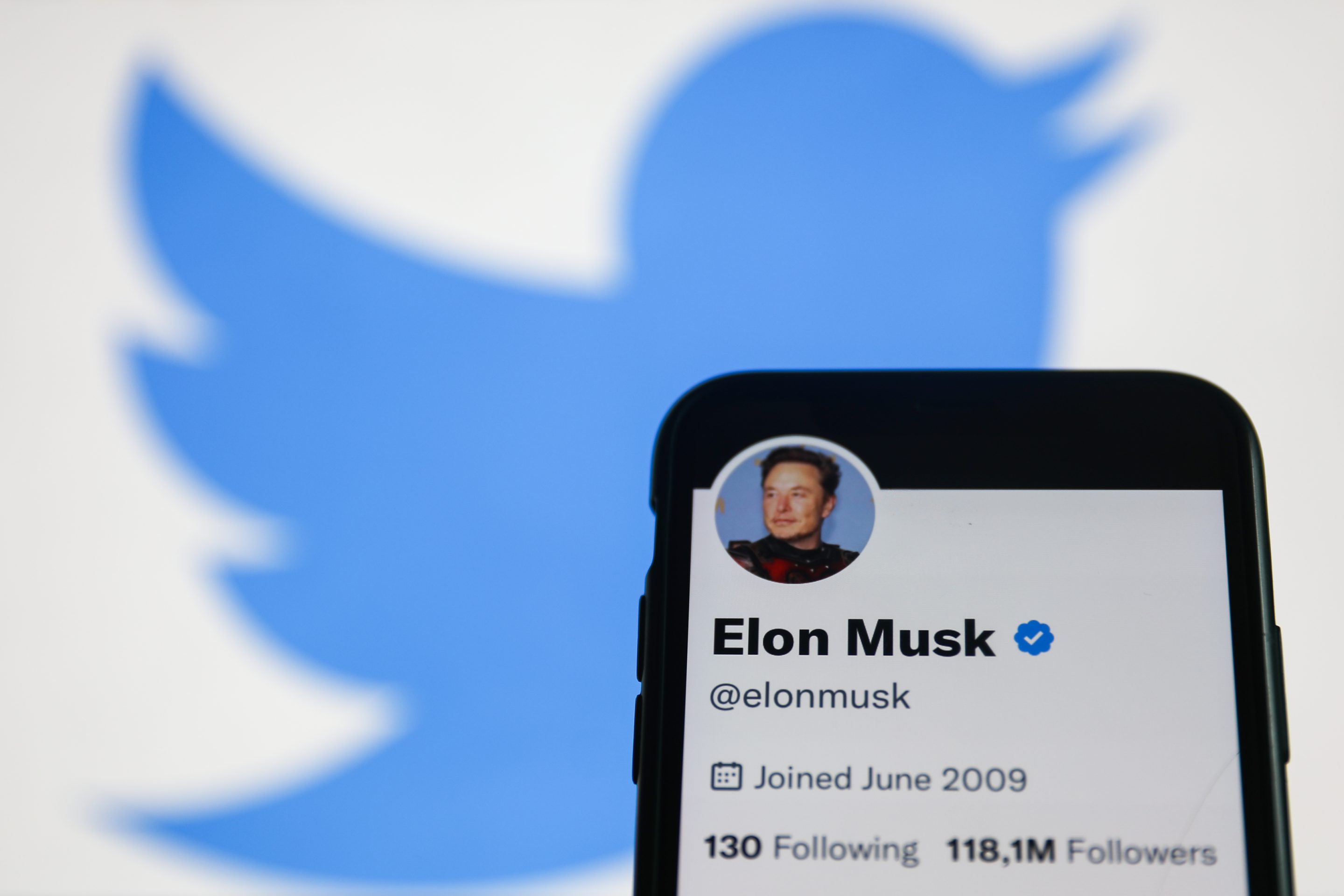 Elon Musk Twitter account displayed on a phone screen and Twitter logo displayed on a screen in the background