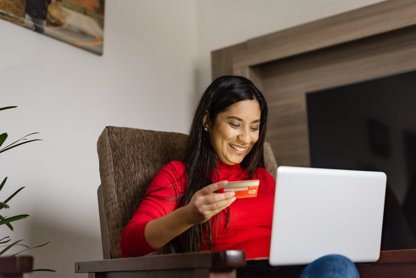 Young woman sitting on the armchair holding a credit card for online shopping on laptop