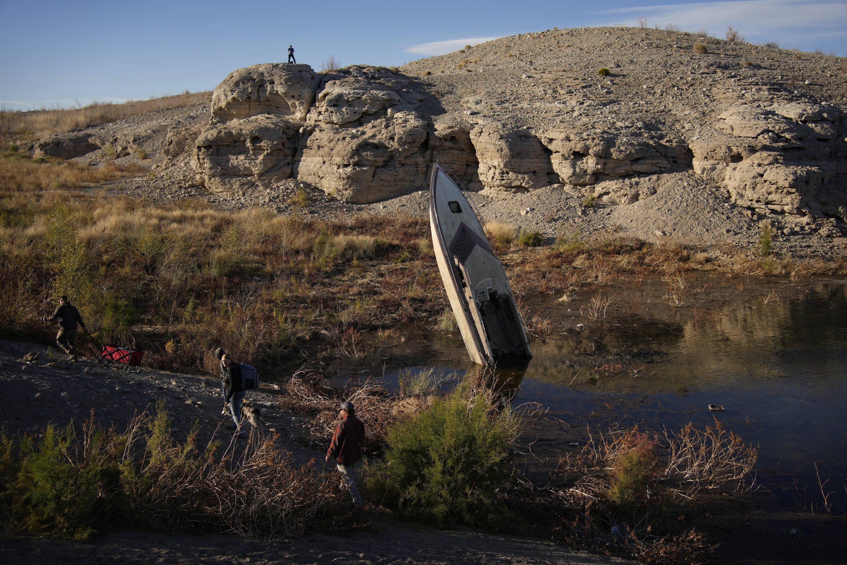 People walk by a formerly sunken boat standing upright into the air with its stern buried in the mud along the shoreline of Lake Mead at the Lake Mead National Recreation Area, on Jan. 27, 2023, near Boulder City, Nev.