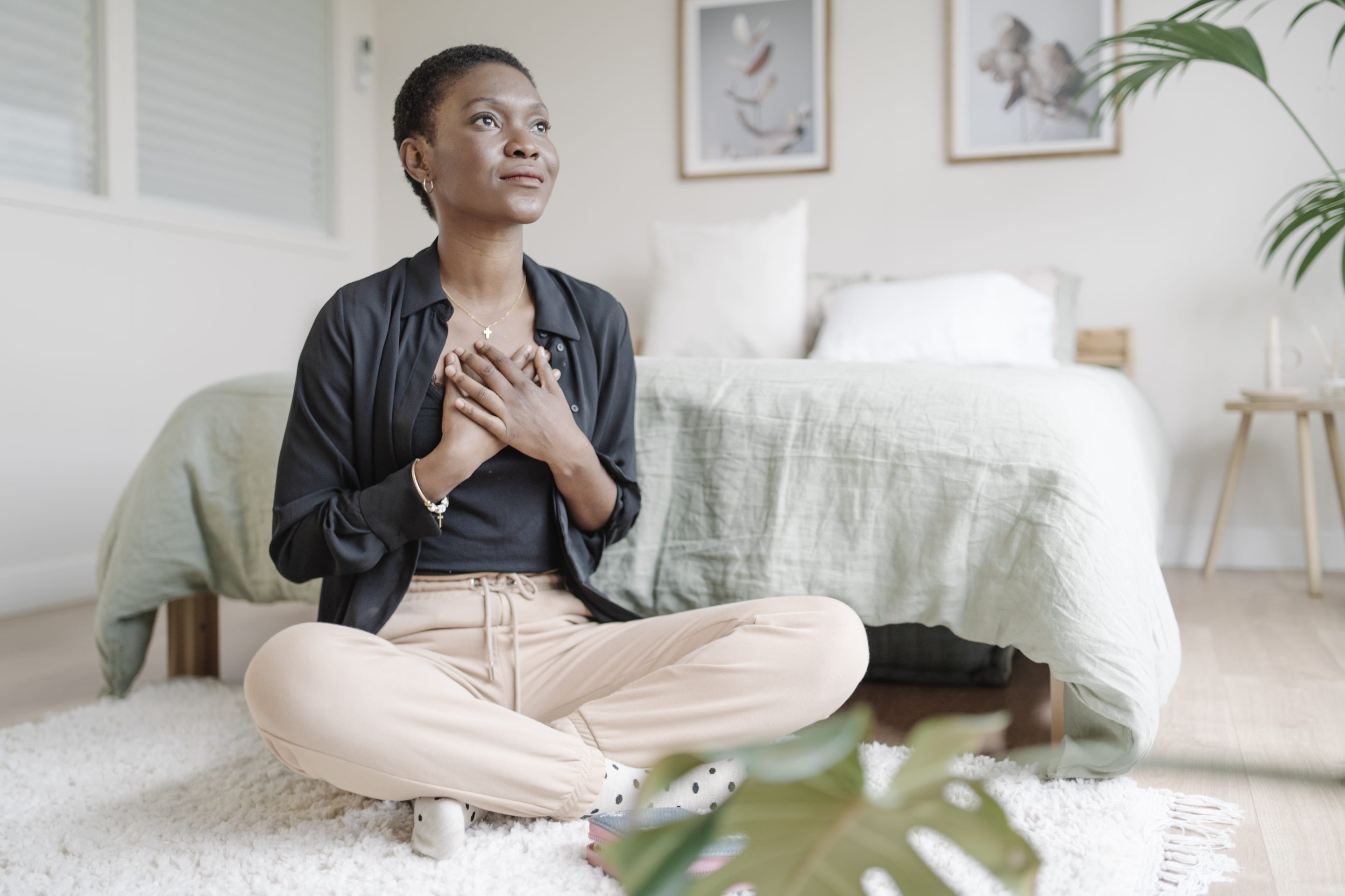 Woman with hand on chest showing gratitude while praying sitting on the floor in her bedroom. Spirituality concept.