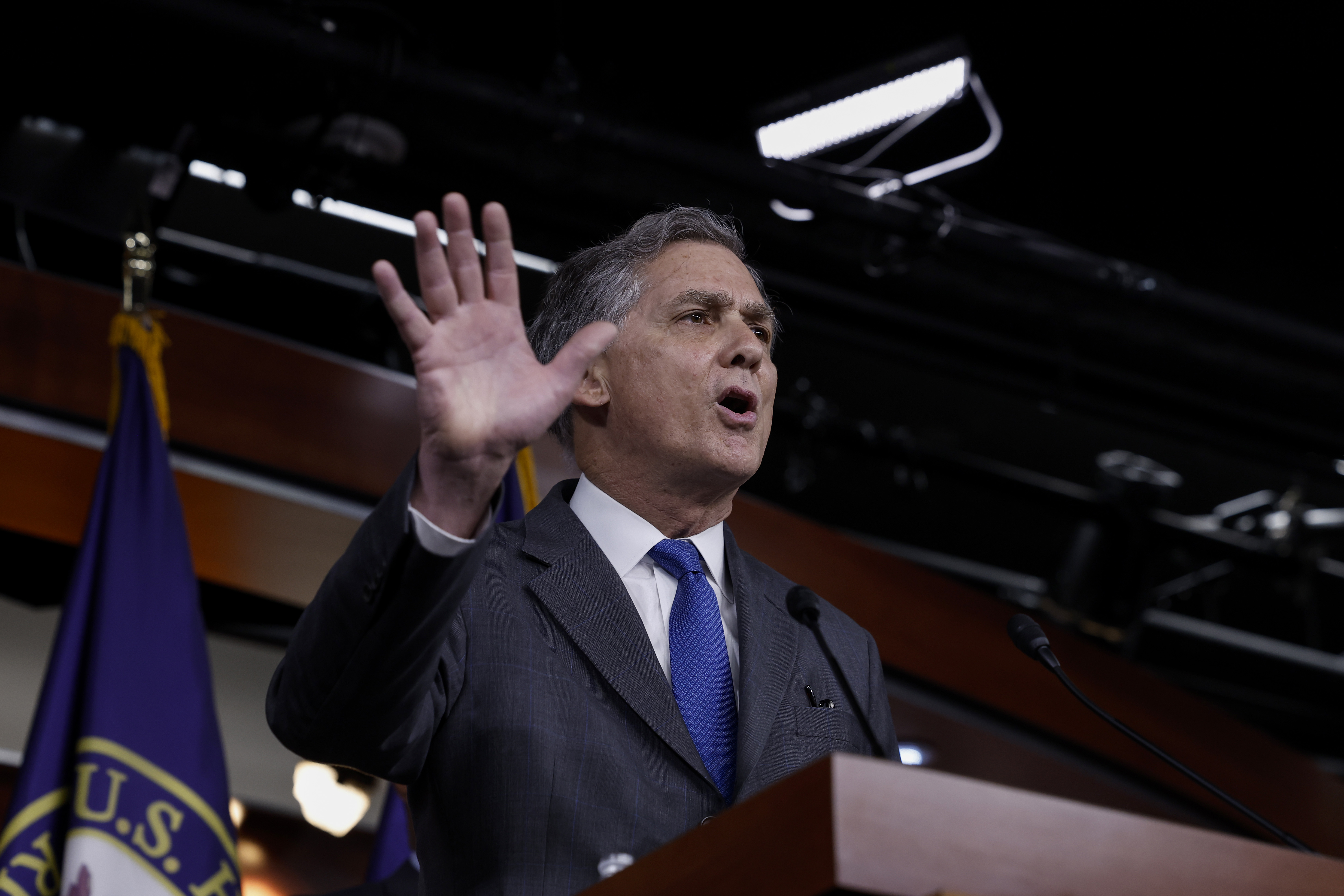 A man dressed in a dark grey suit and blue tie, holds up his right hand towards an audience of reporters, while he stands behind a podium, speaking into a microphone