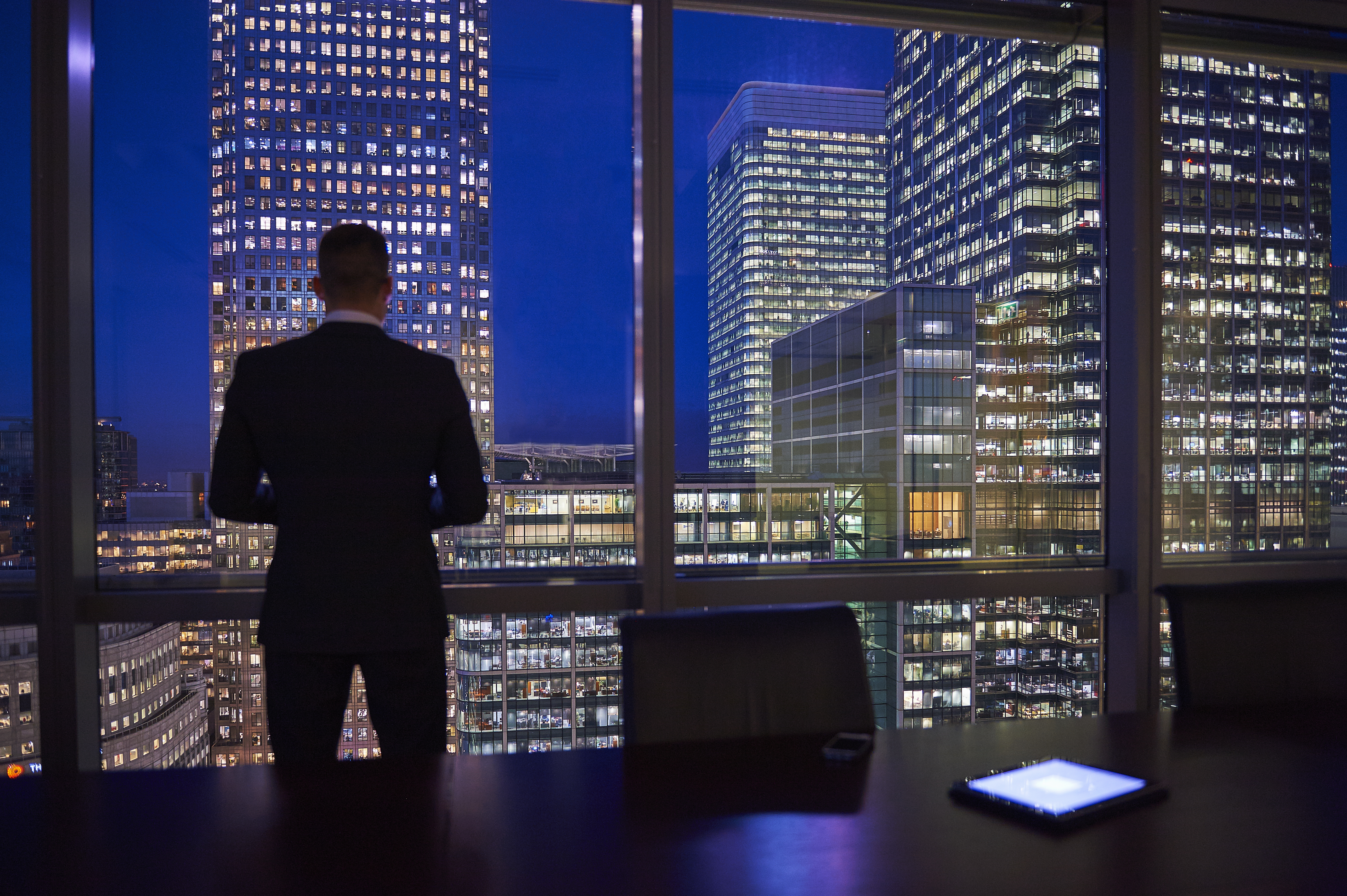 Businessman looking out of a large boardroom window at London's financial district at night