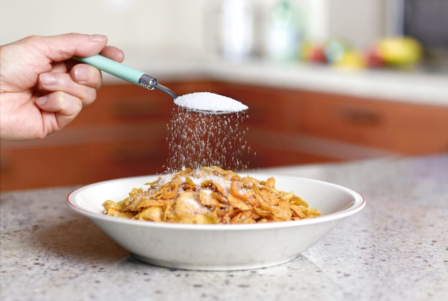 An unseen person sprinkles sugar onto a bowl of cornflakes.