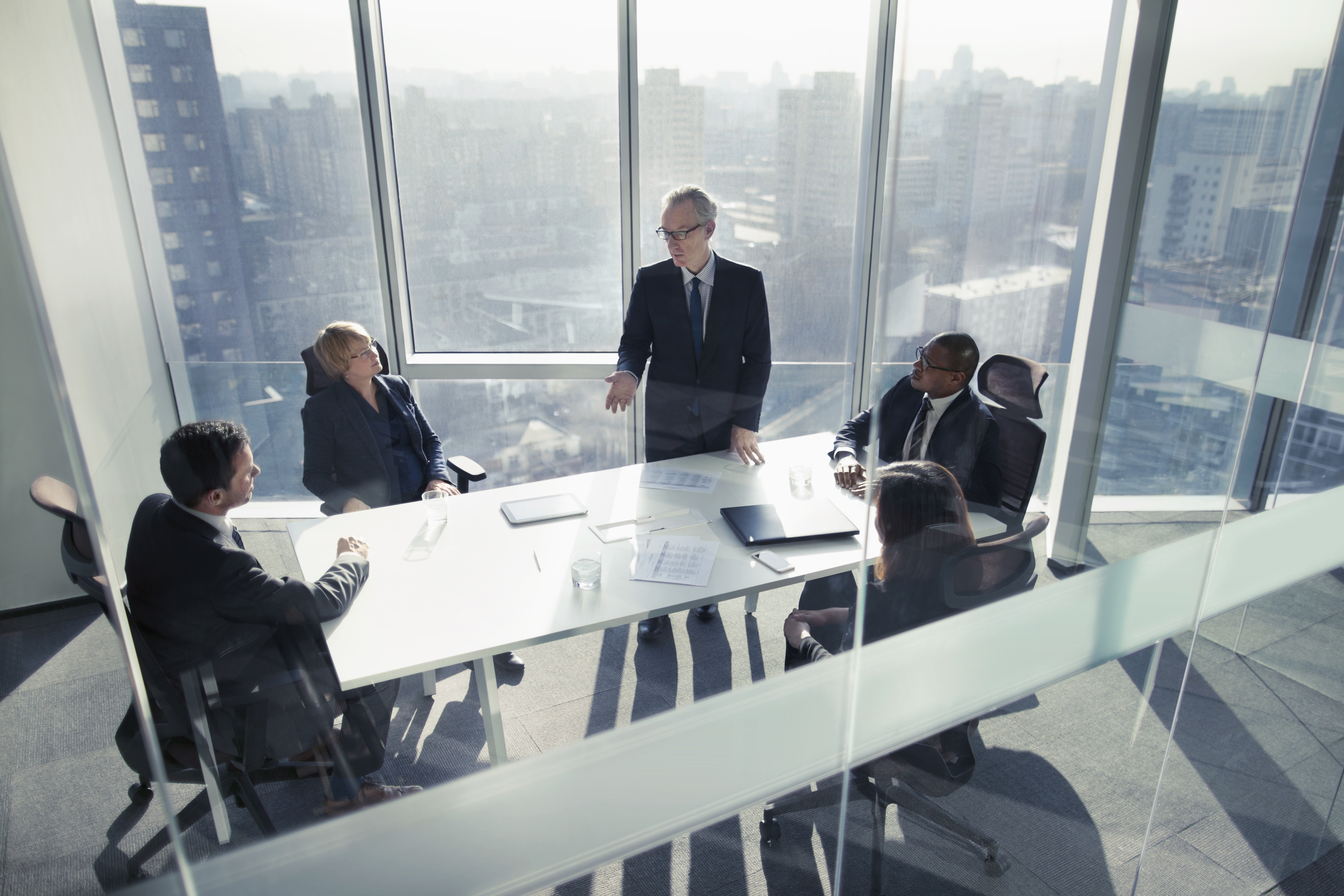 Mature senior adult businessman leader boss CEO executive with group of business people sitting together in conference meeting room in contemporary modern office bright sunny daylight talking discussing planning organizing strategy well dressed suit and tie teamwork cooperation diversity multi-ethnic problems solutions unity cityscape downtown urban