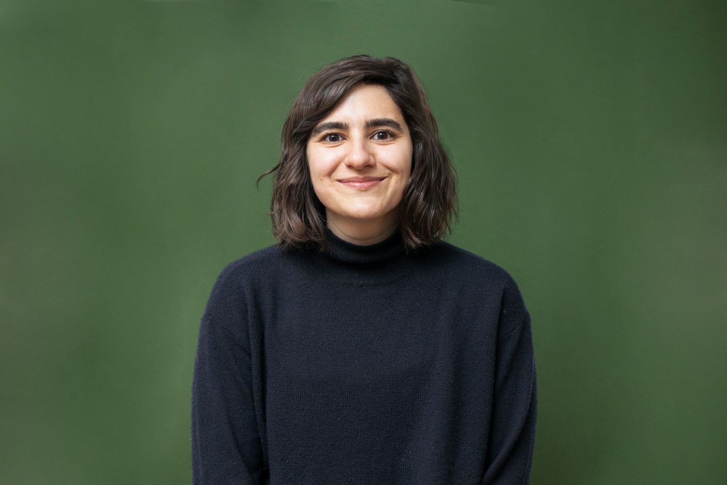 A young woman wearing a black sweater smiles as she poses in front of a dark green background