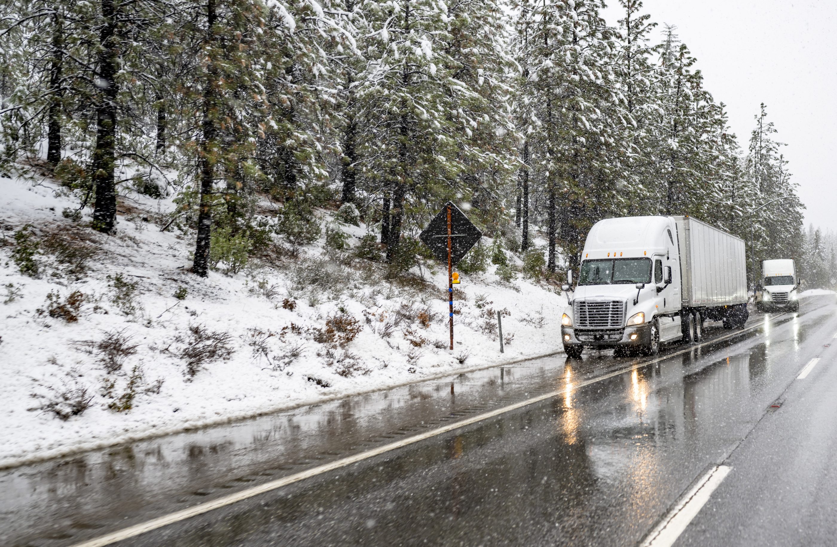 Professional industrial logistic two big rig semi trucks with turned on headlight loaded semi trailers standing on the wet highway road shoulder waiting for the end of the winter snowing storm.