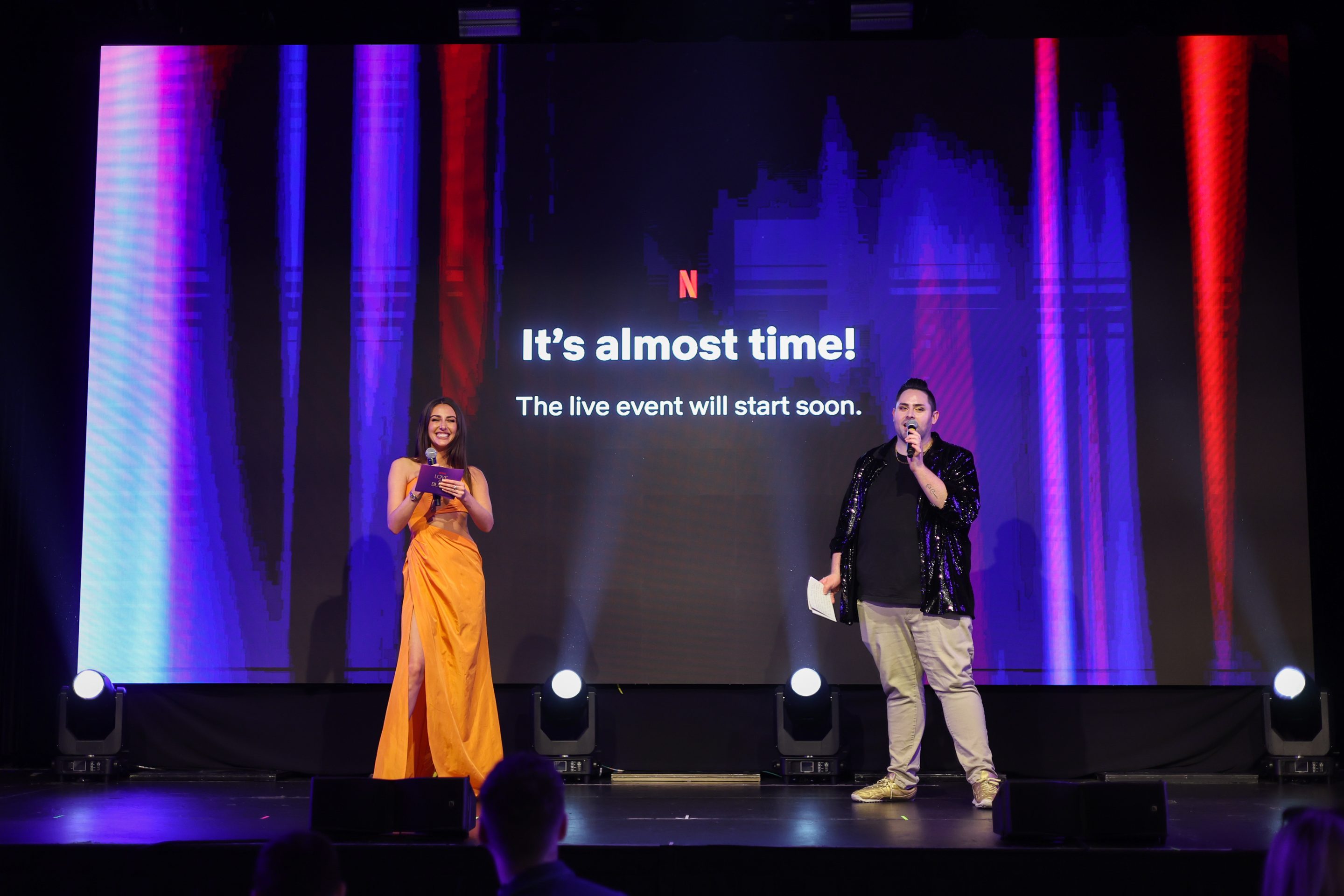 Two hosts stand in front of the Netflix waiting room screen for the Love is Blind live reunion special.
