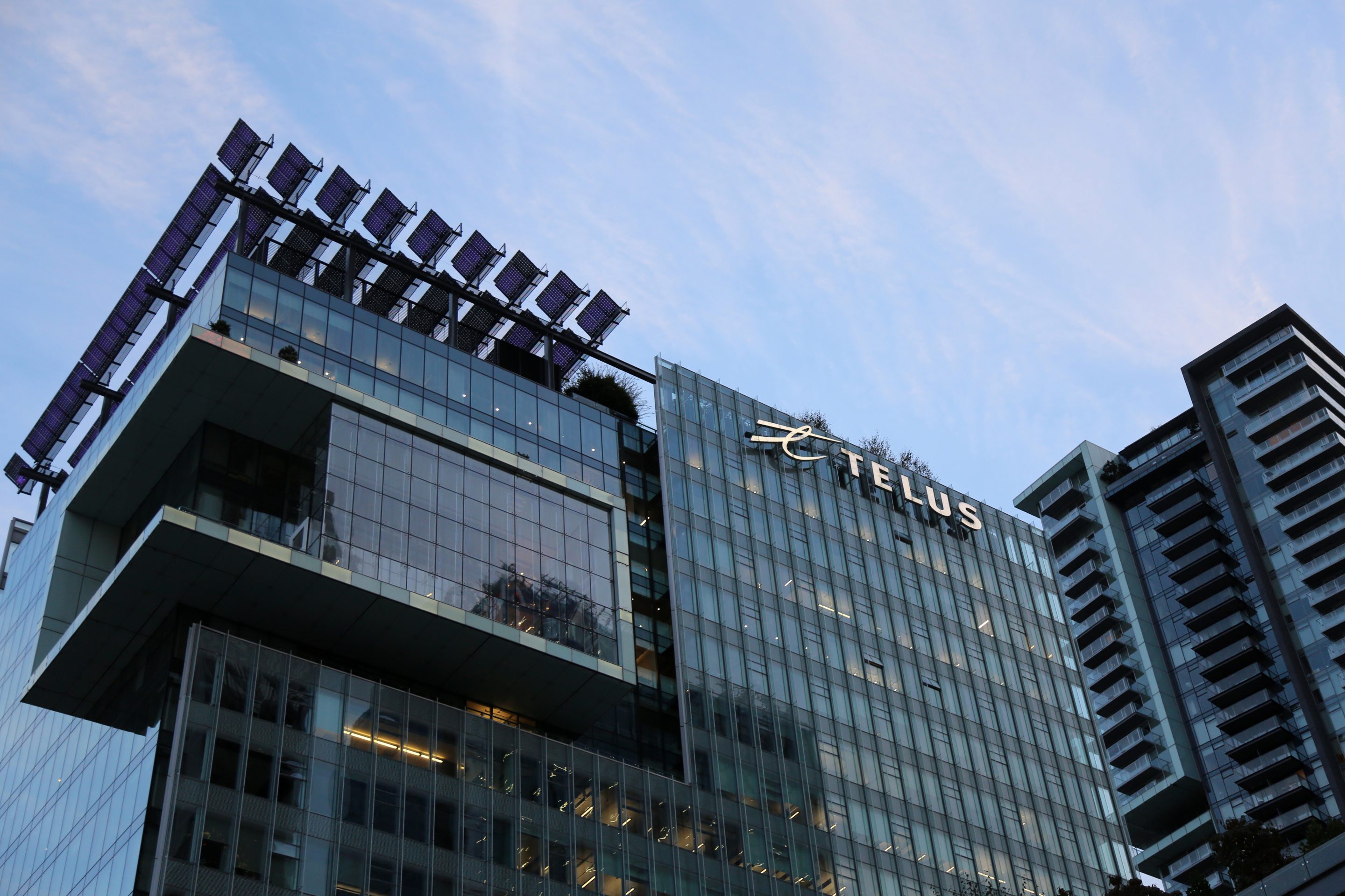 A glass and steel building towers against the blue sky. A sign reading "Telus" is mounted to the side of the building
