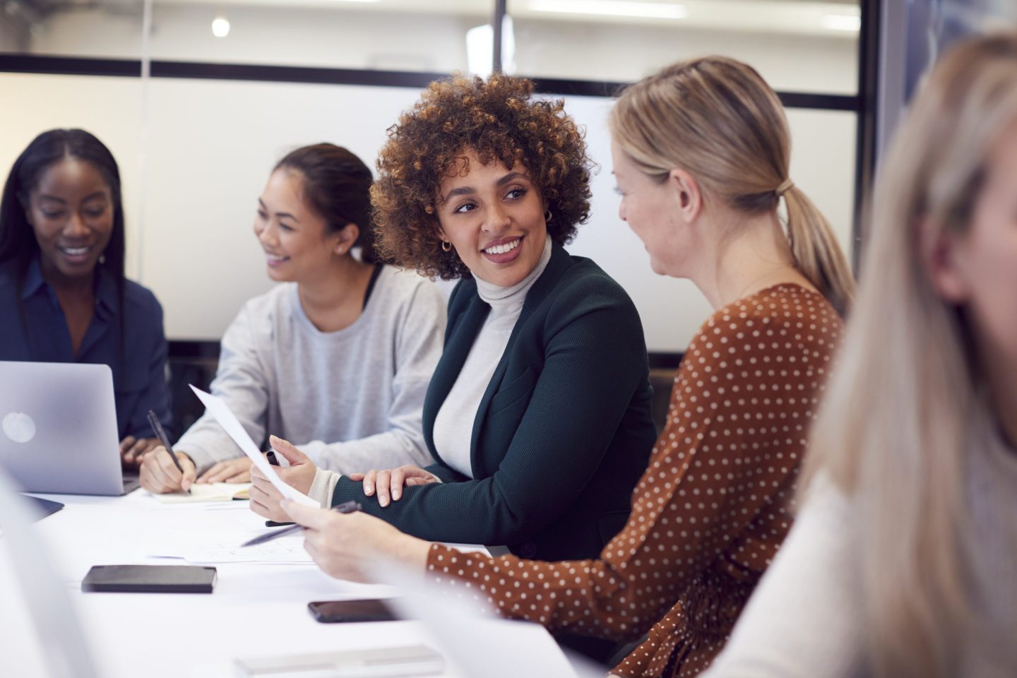 A group of women sit around a conference room table and discuss amongst themselves.