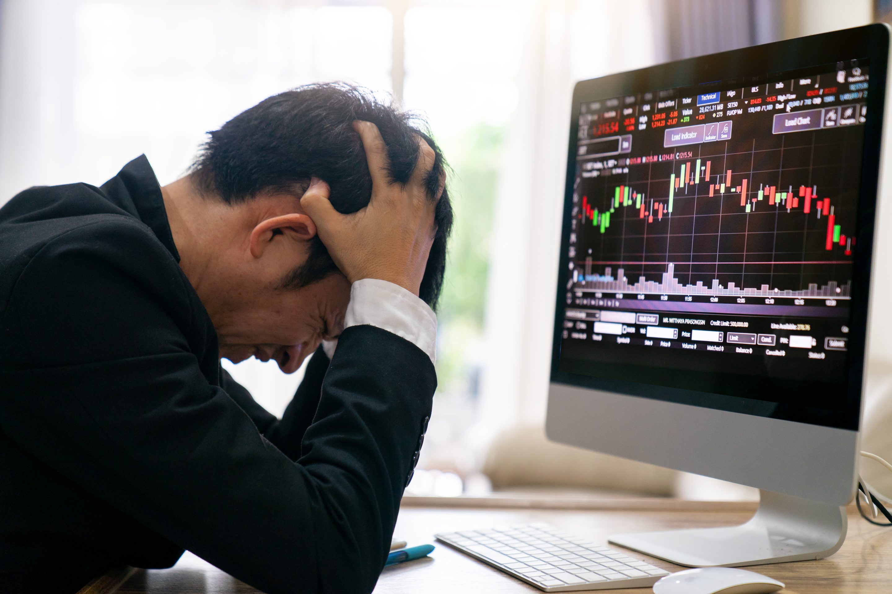 Stressed businessman, head in hands, sits before computer screen displaying stock market investments.