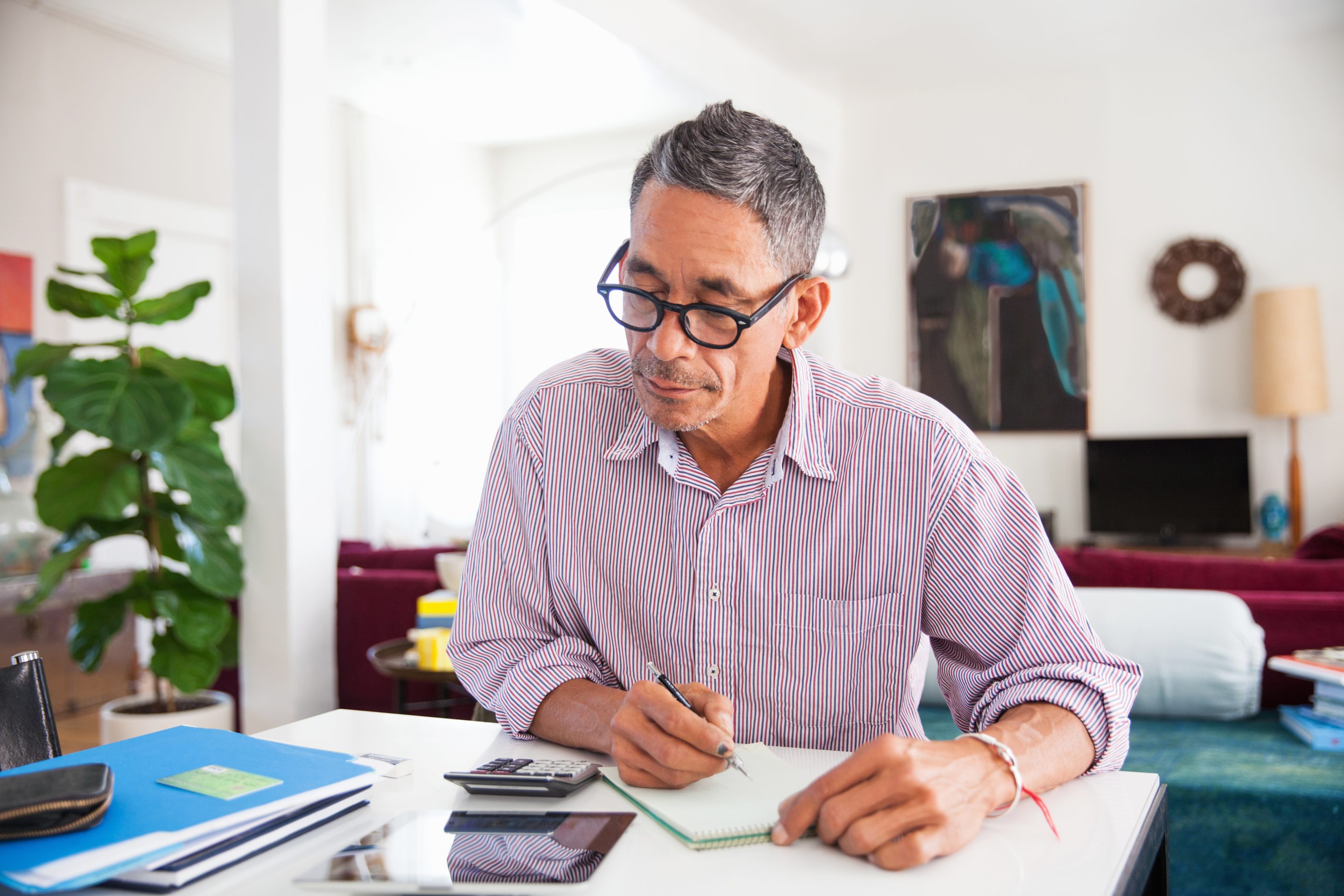 Stock image of a man sitting at a desk looking at a calculator and writing notes on a notepad.