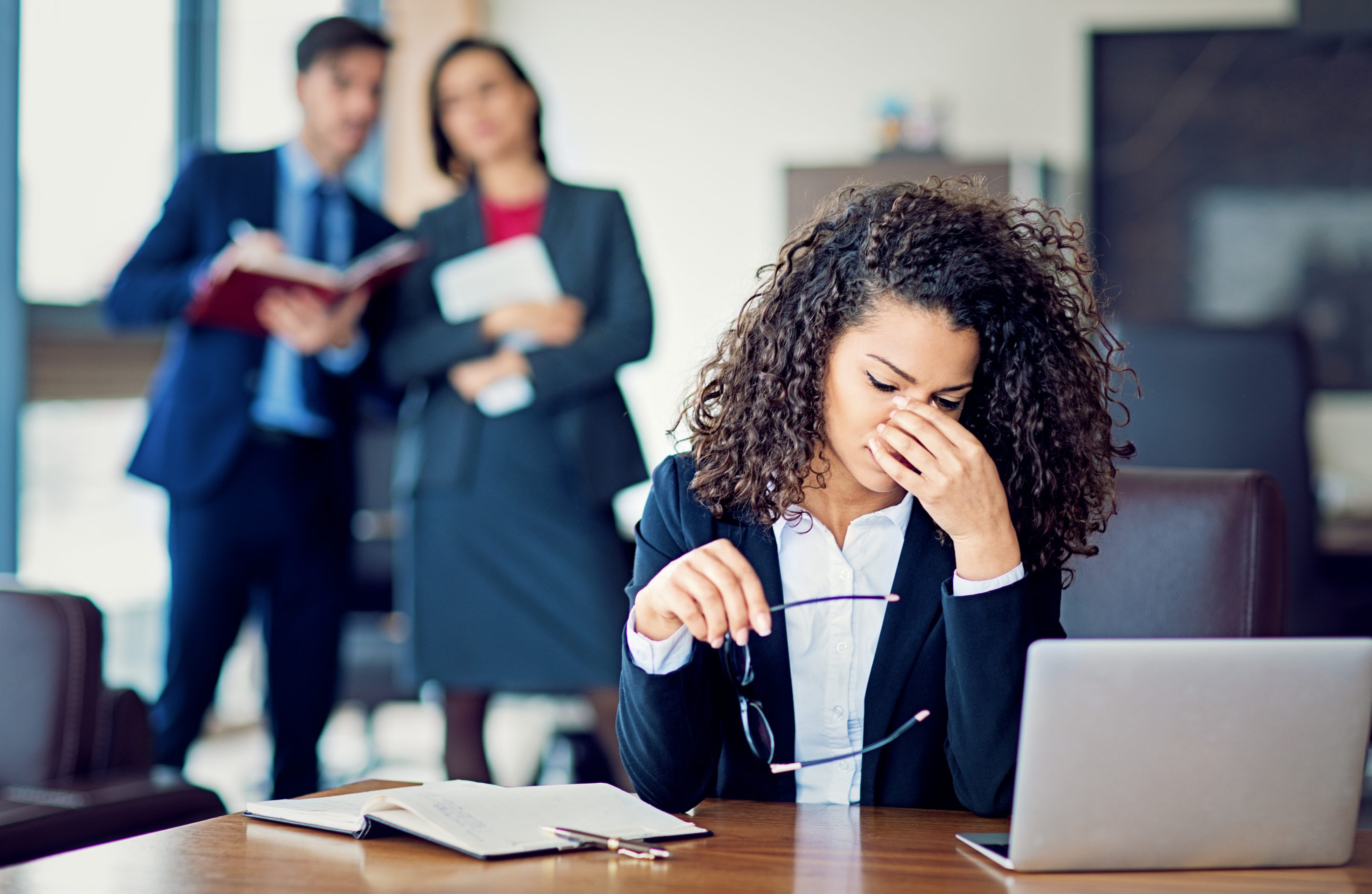 Stock photo of a businesswoman looking stressed while sitting at her desk, as two colleagues look on.