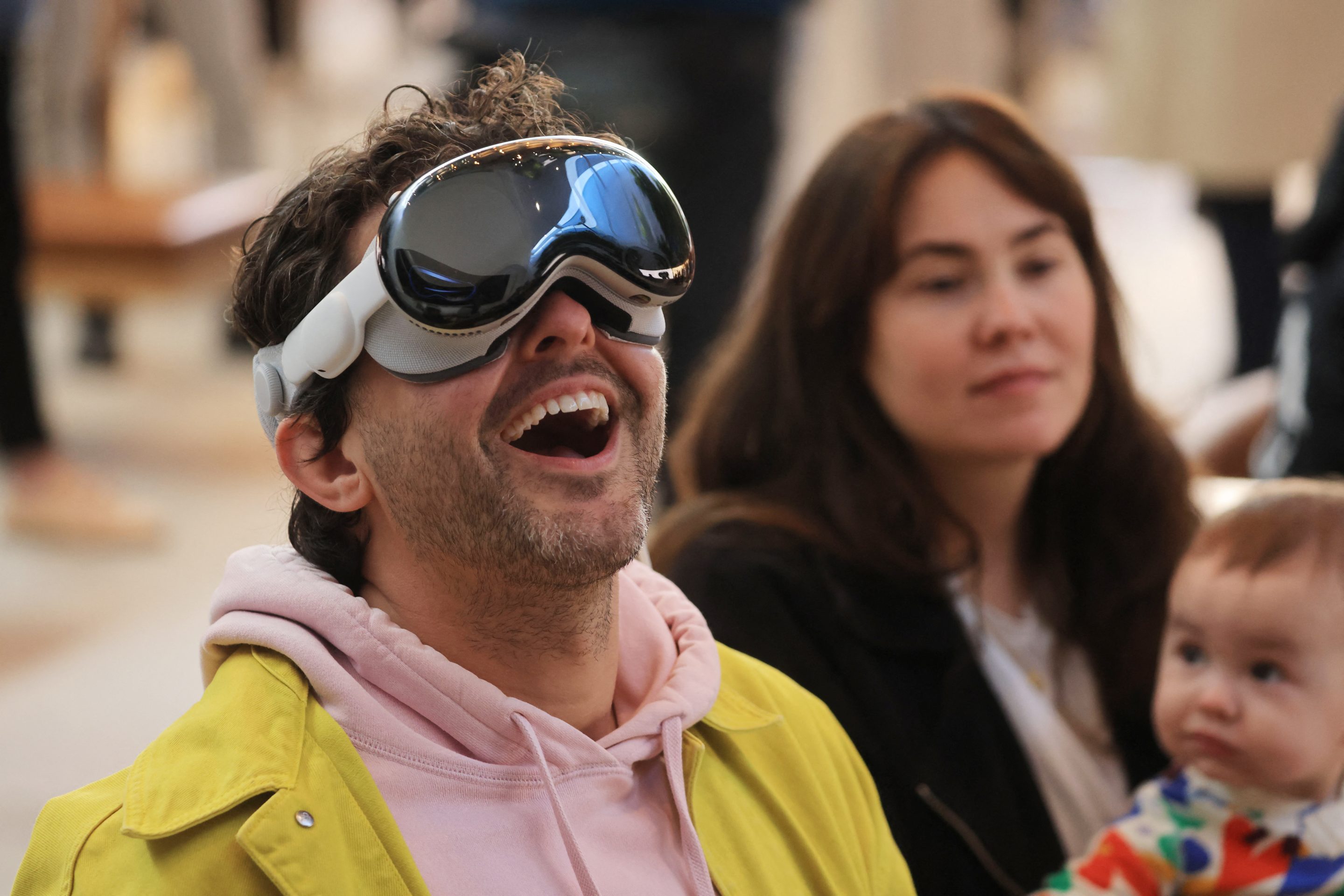 A man in a pink hoodie and yellow jacket is wearing the Apple Vision Pro, looking up, and smiling in awe.