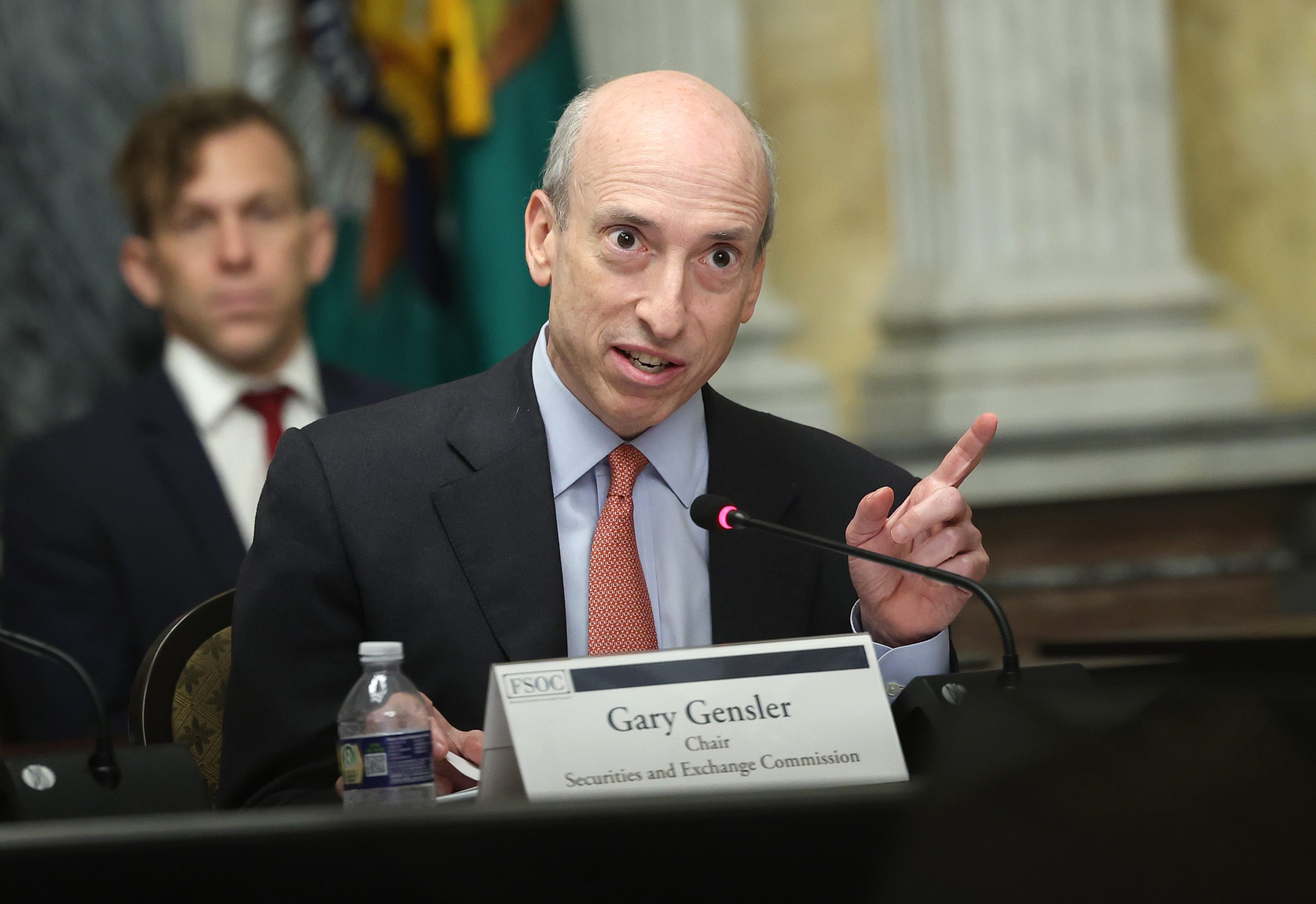 A man in a dark suit, light blue shirt, and red tie sits behind a table with a name card in front reading "Gary Gensler"
