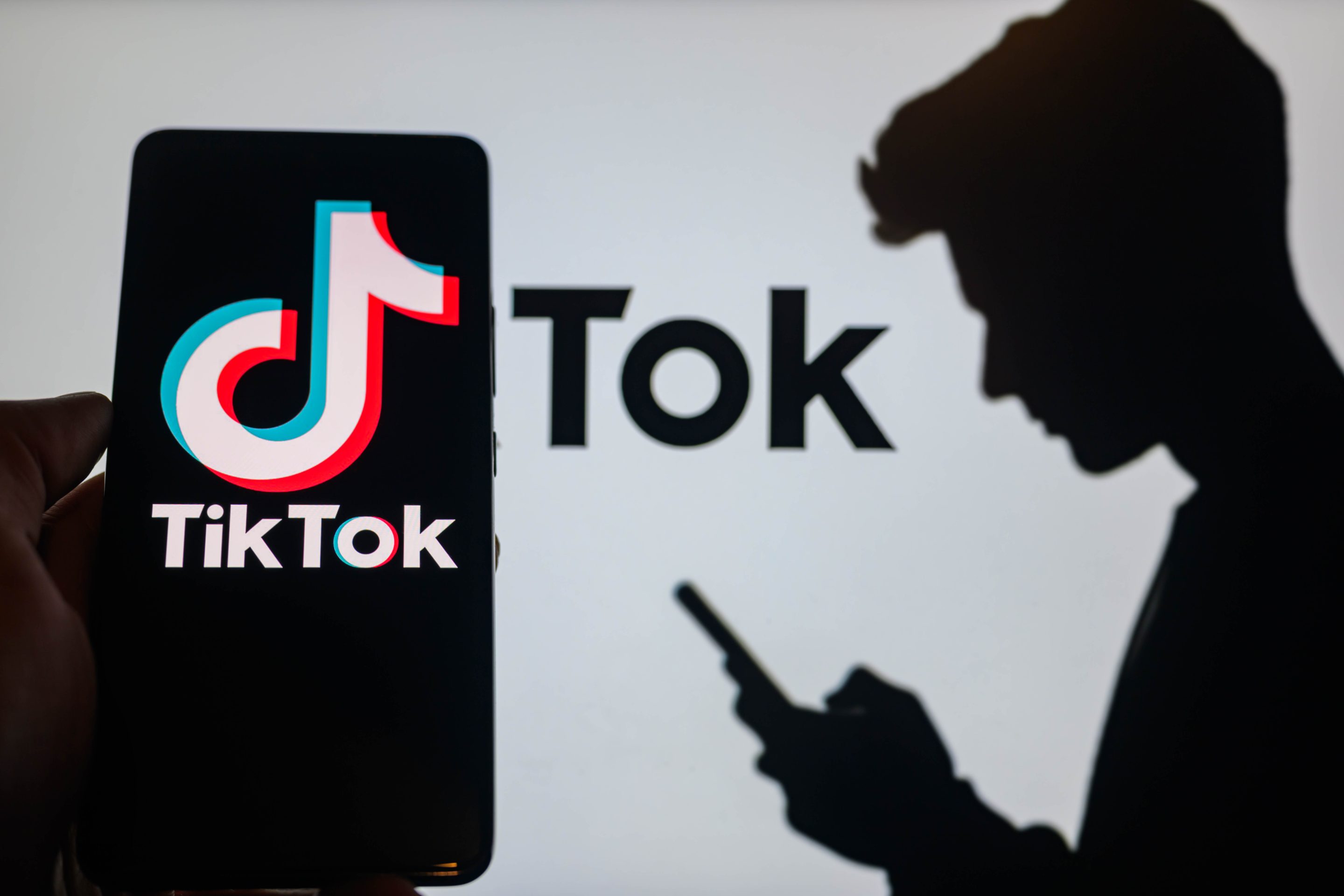 The TikTok logo is being displayed on a smartphone with TikTok visible in the background