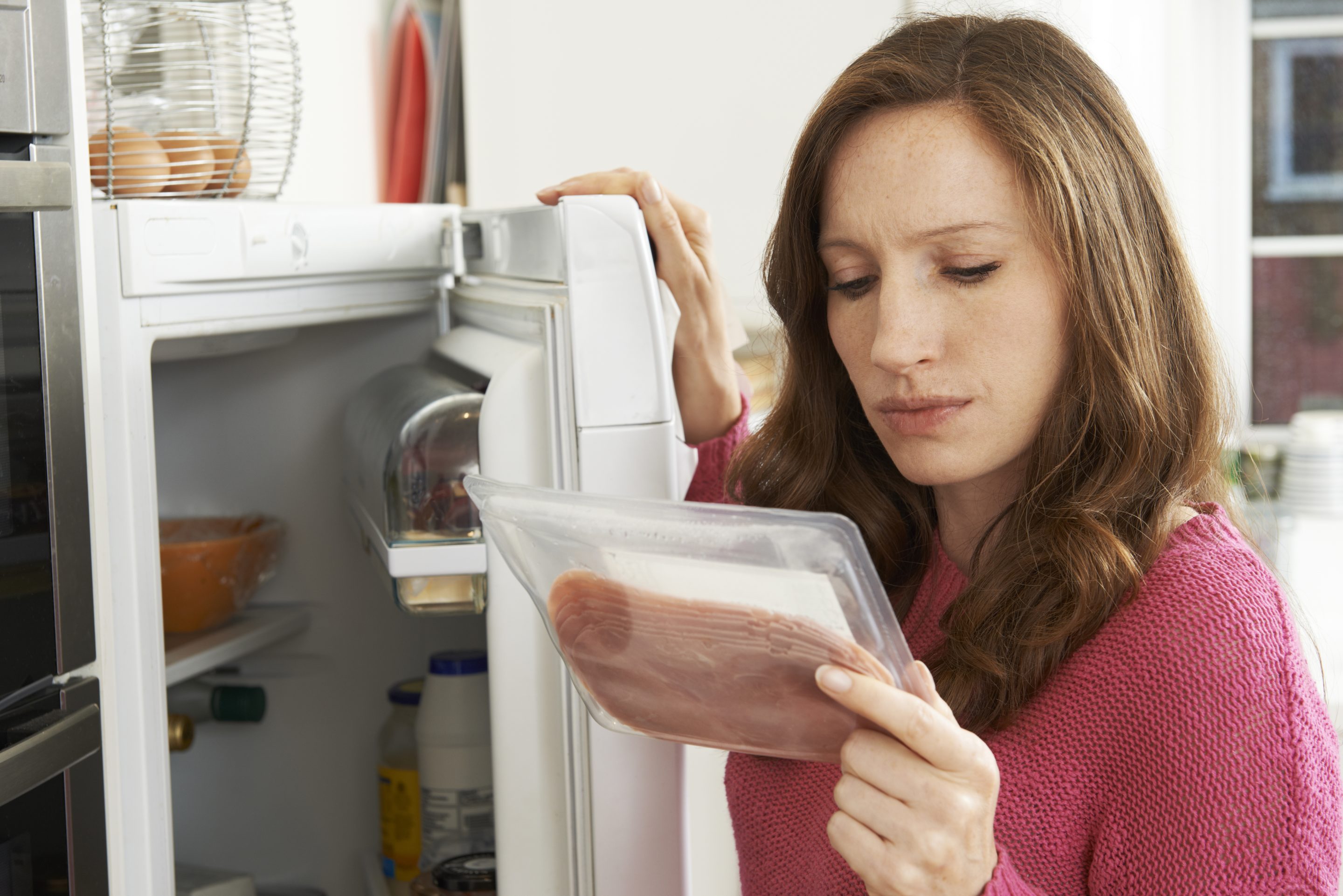 A woman stands next to her open fridge and looks down at a package of sliced deli meat.