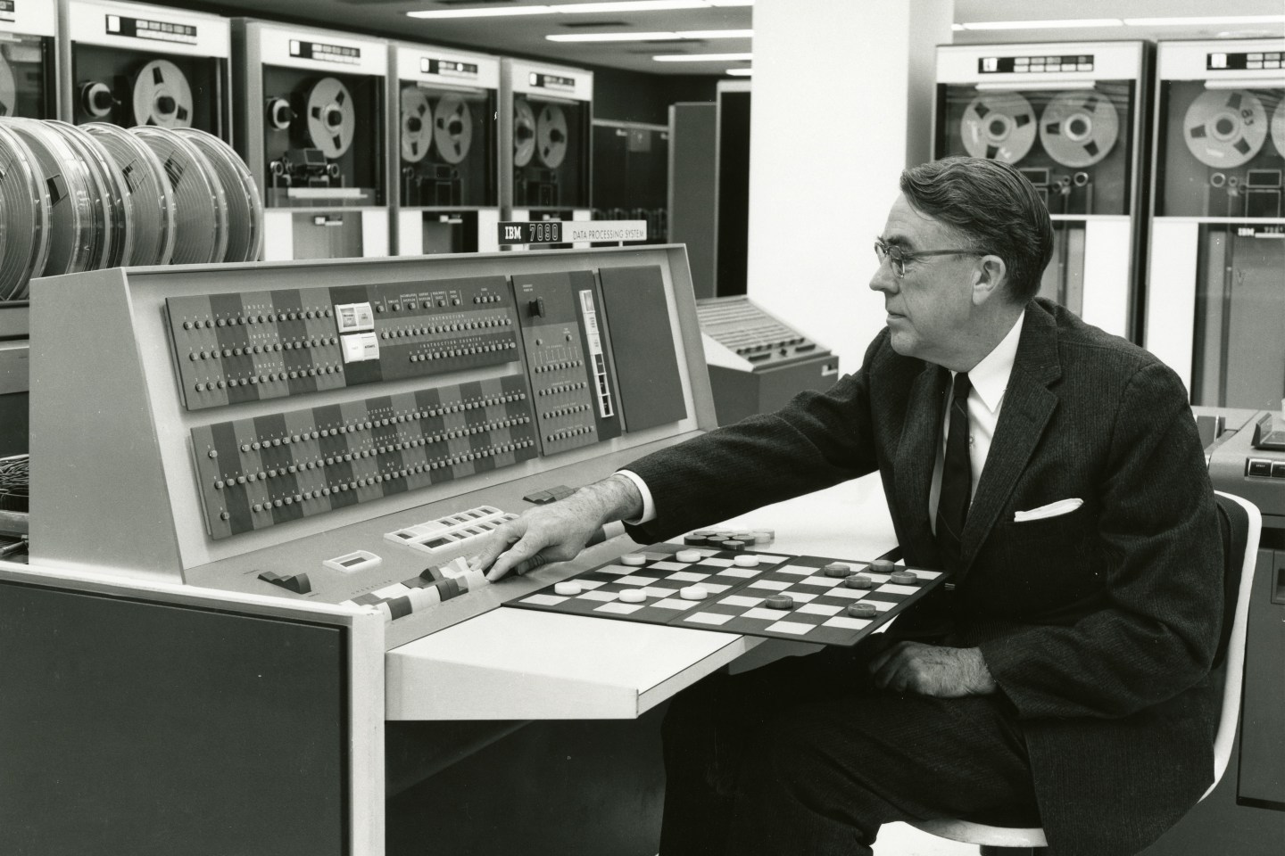 A middle-aged man in a suit sits in front of a checker board, working a computer's control panel.
