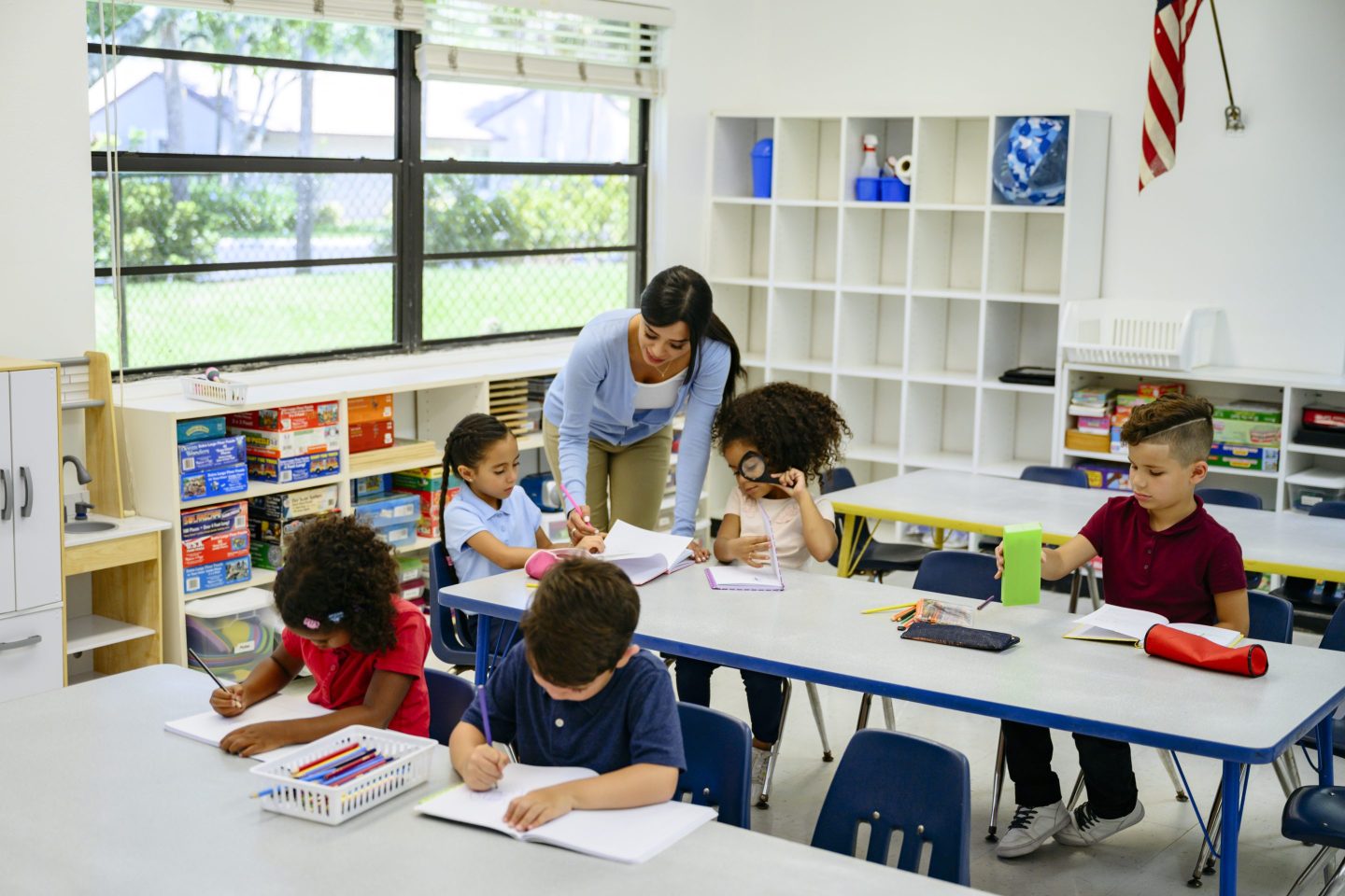 A young woman teacher is bending over at a table at school where young students are working