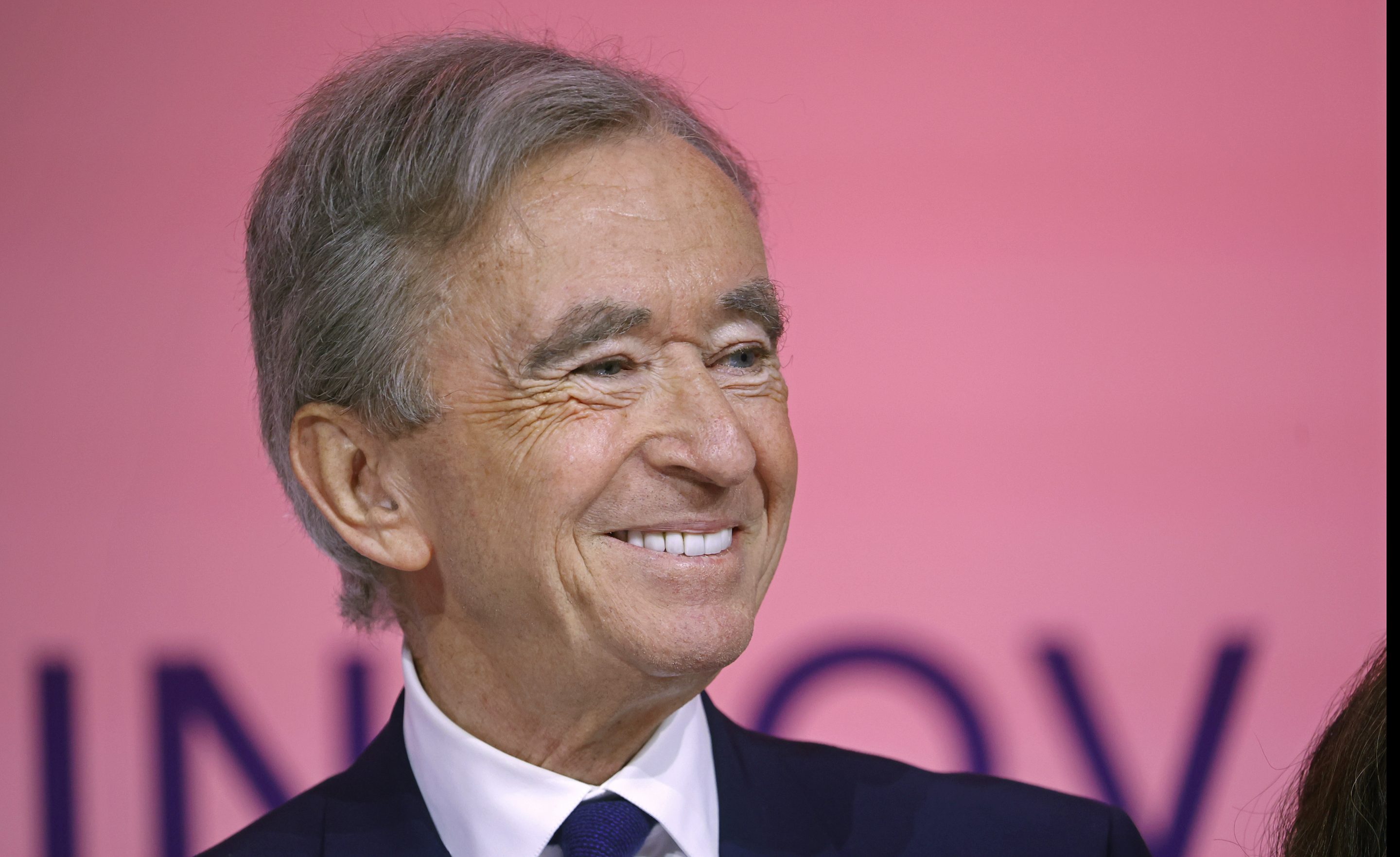 Bernard Arnault smiles to his right and is in front of a pink background.