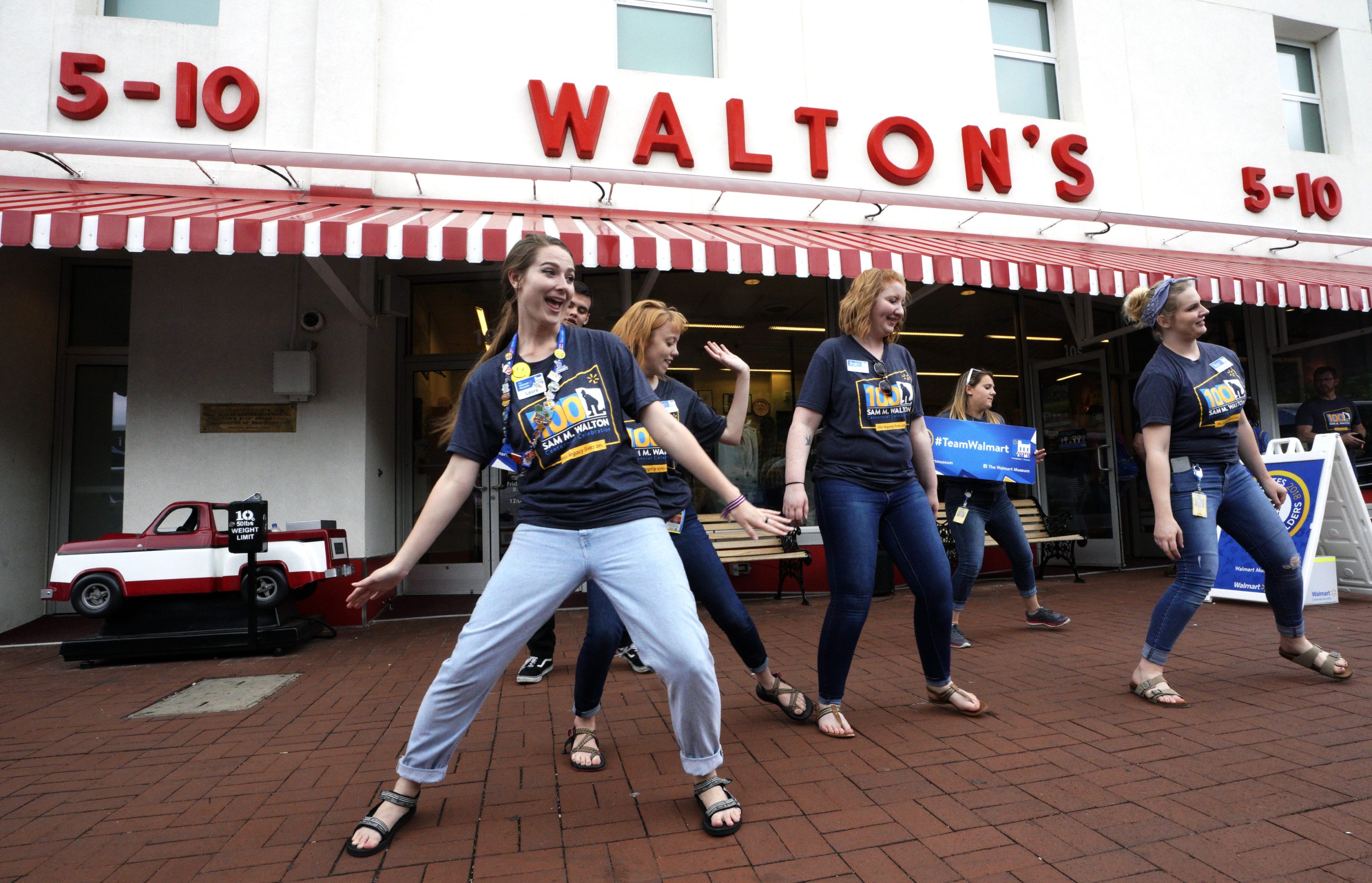 A group of young women dance in front of a white-and-red storefront with a "Walton's" sign across it.