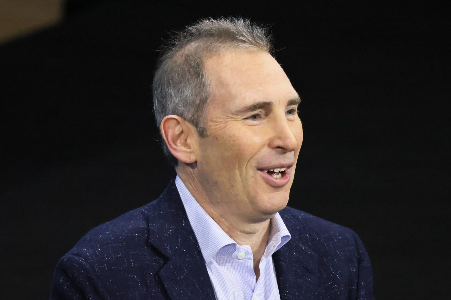 Andy Jassy in a suit and no tie speaks in front of a black background.